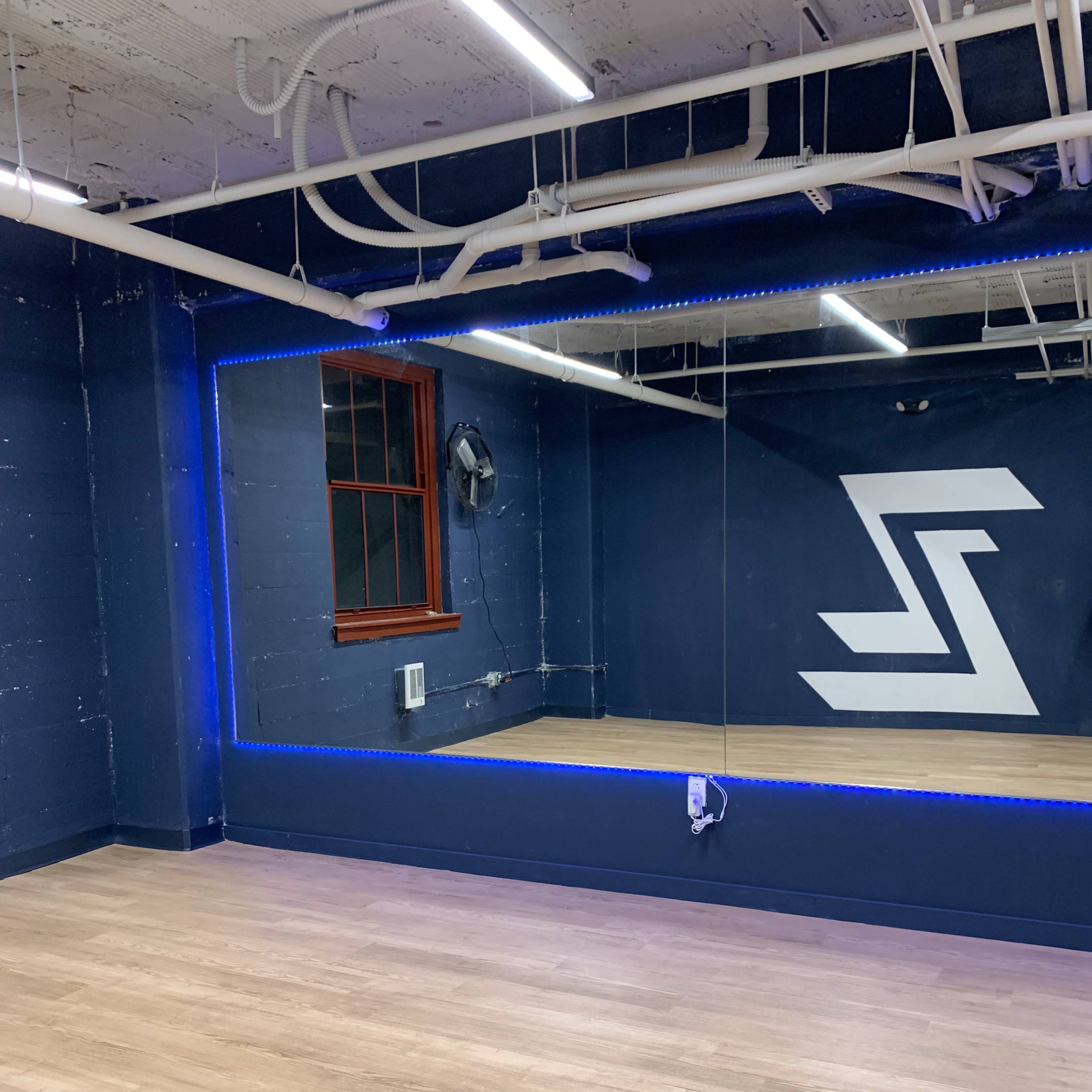 A dance studio with wooden flooring, a large mirror, blue accent lighting, and a wall featuring a white logo.