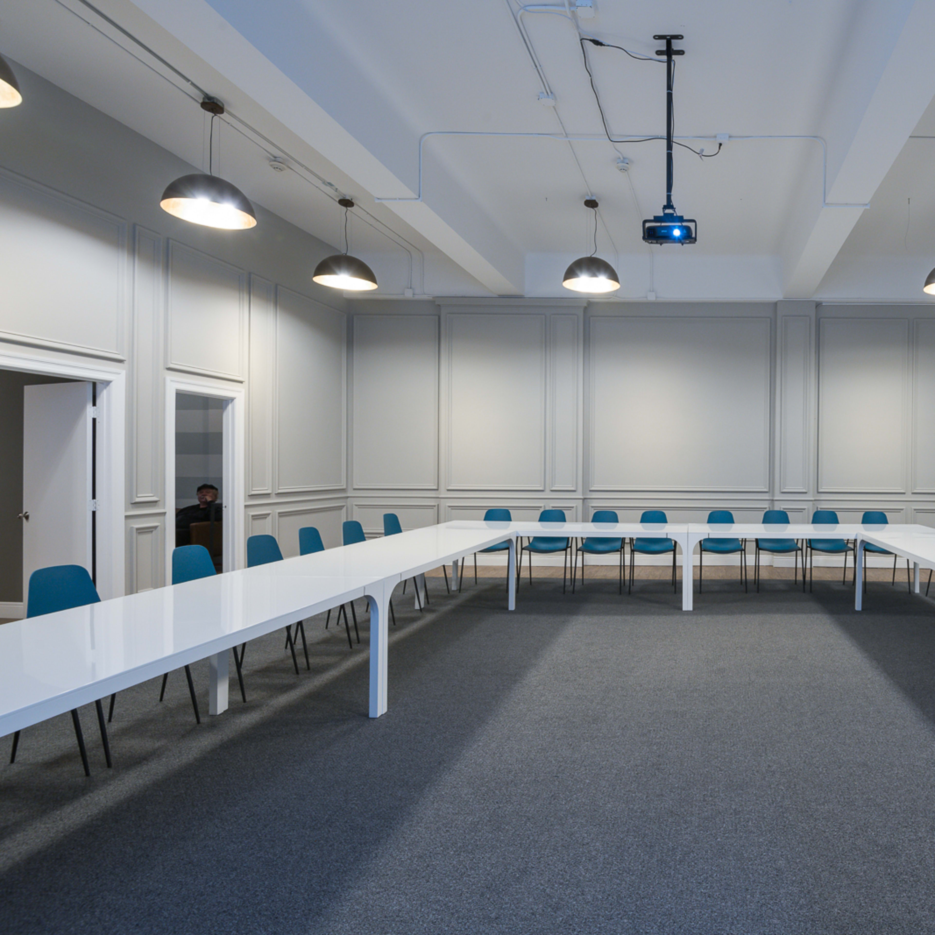 A spacious meeting room features a large white table arranged in a U-shape with blue chairs around it, under industrial-style lighting.