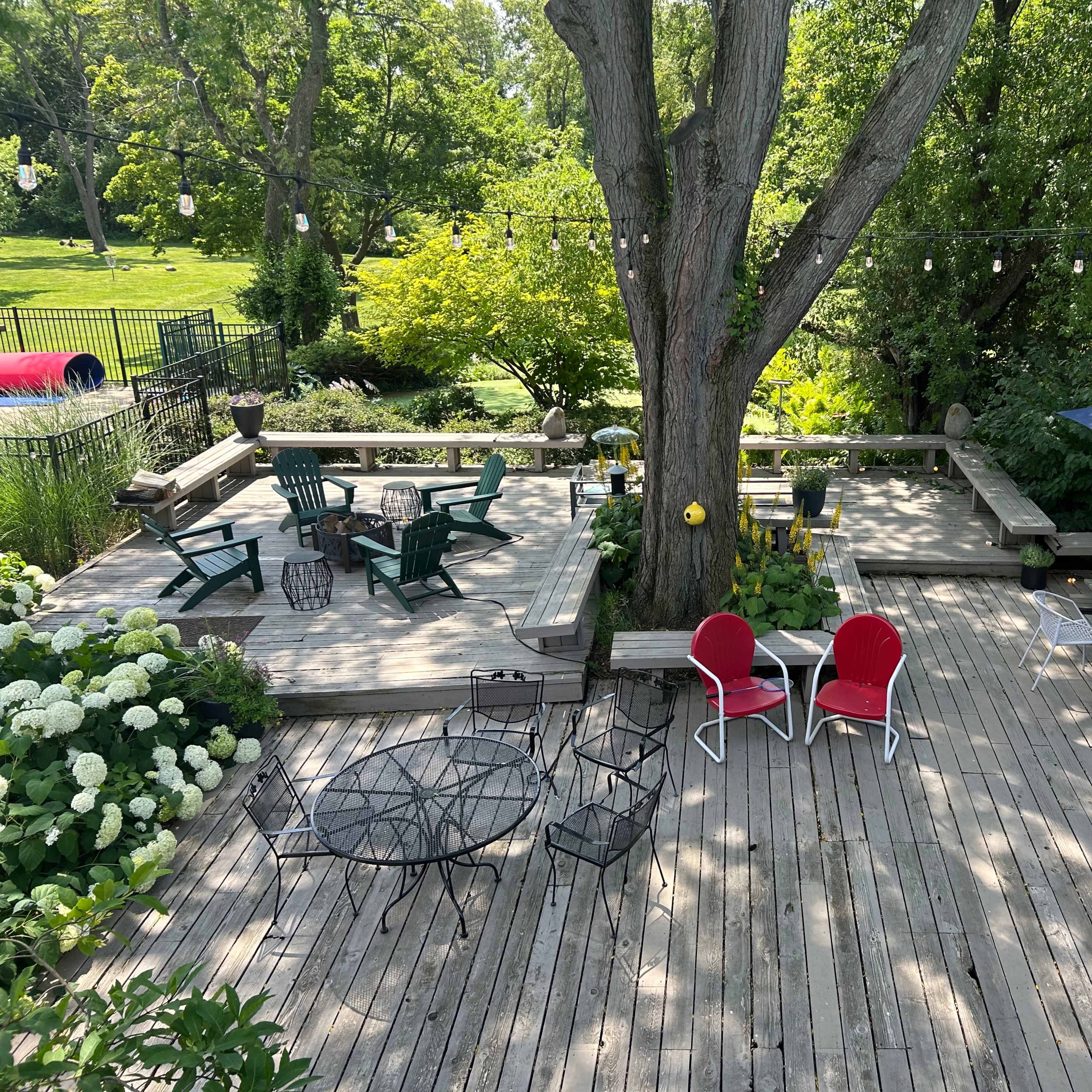 A wooden deck features various seating arrangements, including chairs and tables, surrounded by greenery and shaded by a large tree.