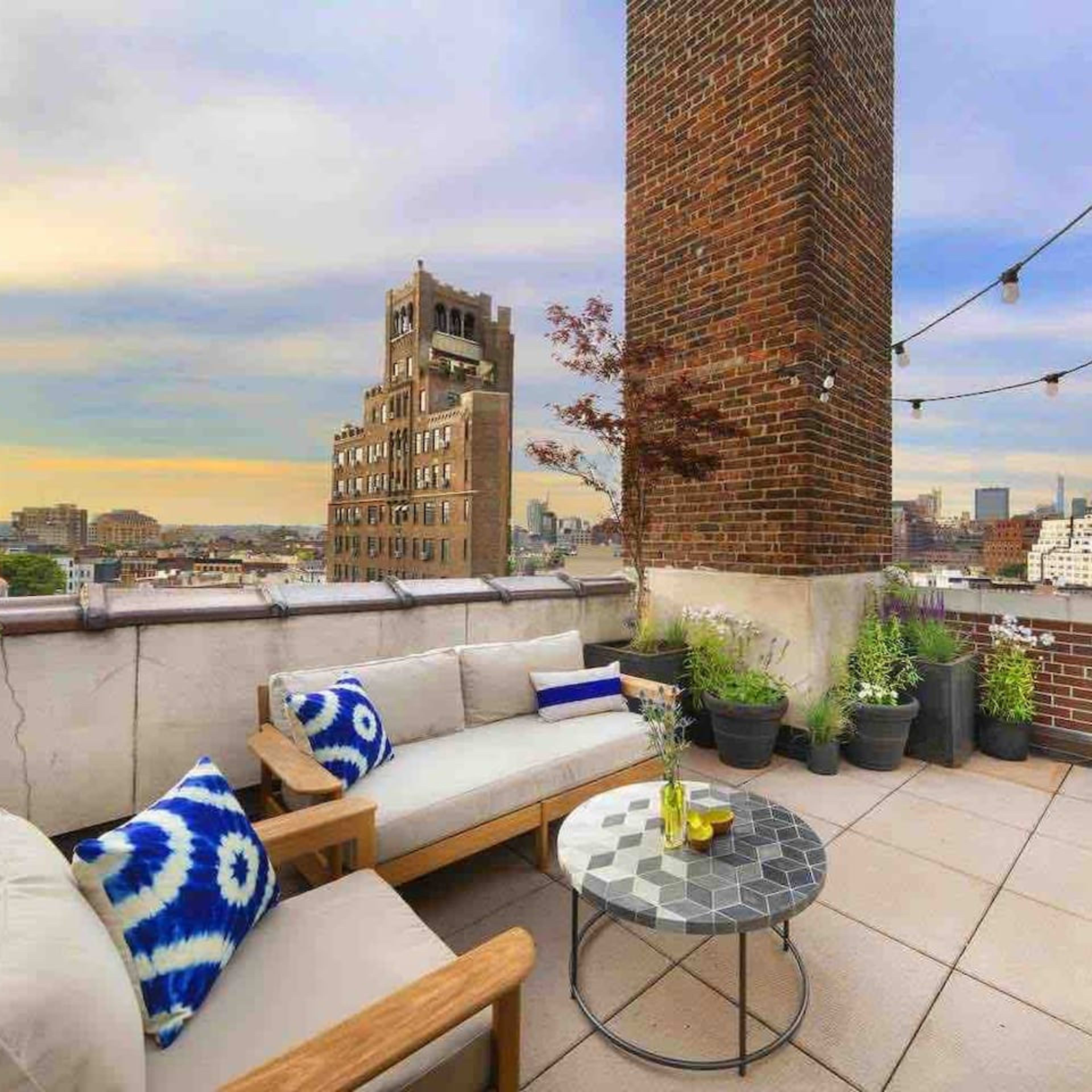 A rooftop terrace features a seating area with a coffee table, surrounded by plants and a view of city buildings.