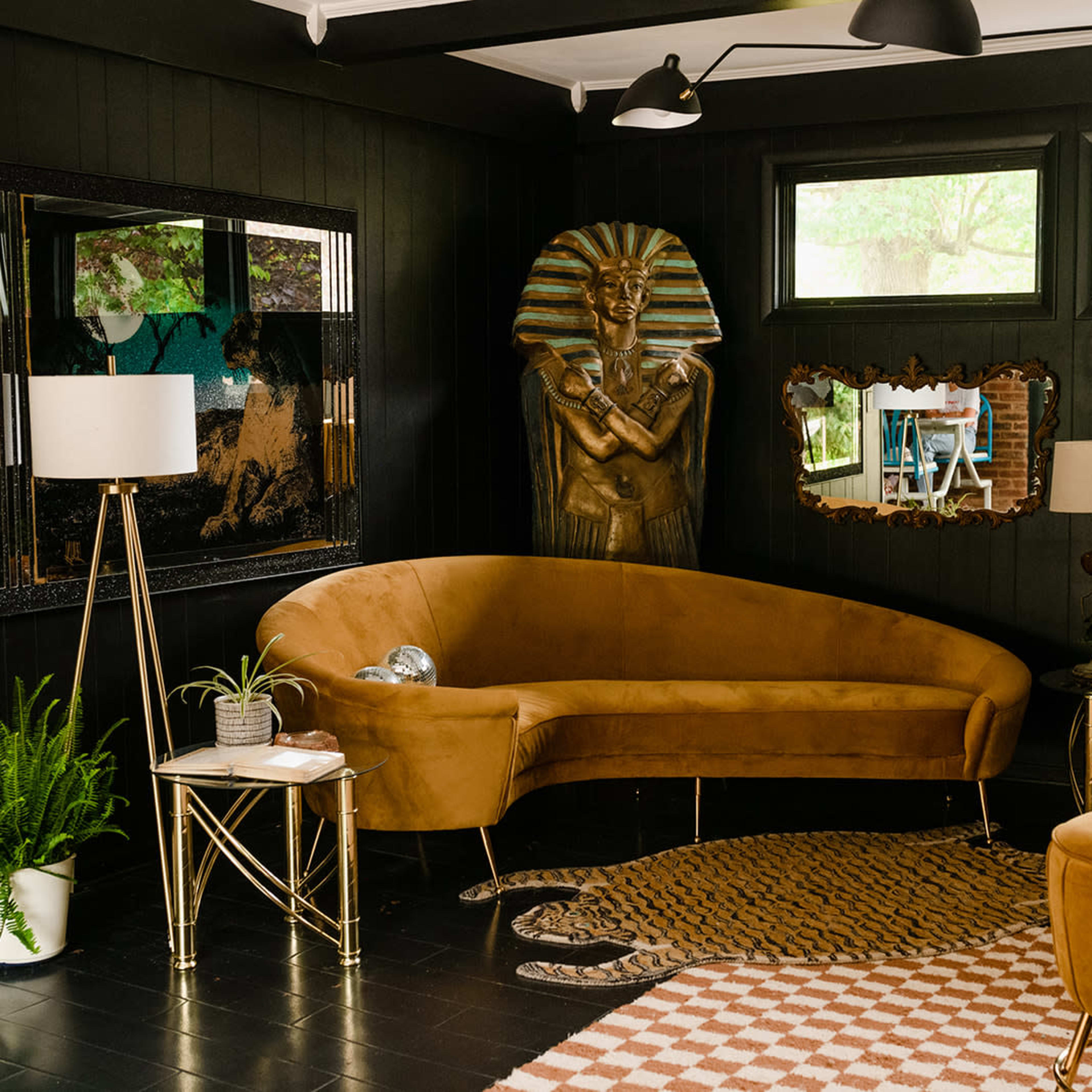 A stylish living room featuring a curved brown sofa, gold-accented furniture, and a statue of an Egyptian pharaoh in a dark-paneled space.