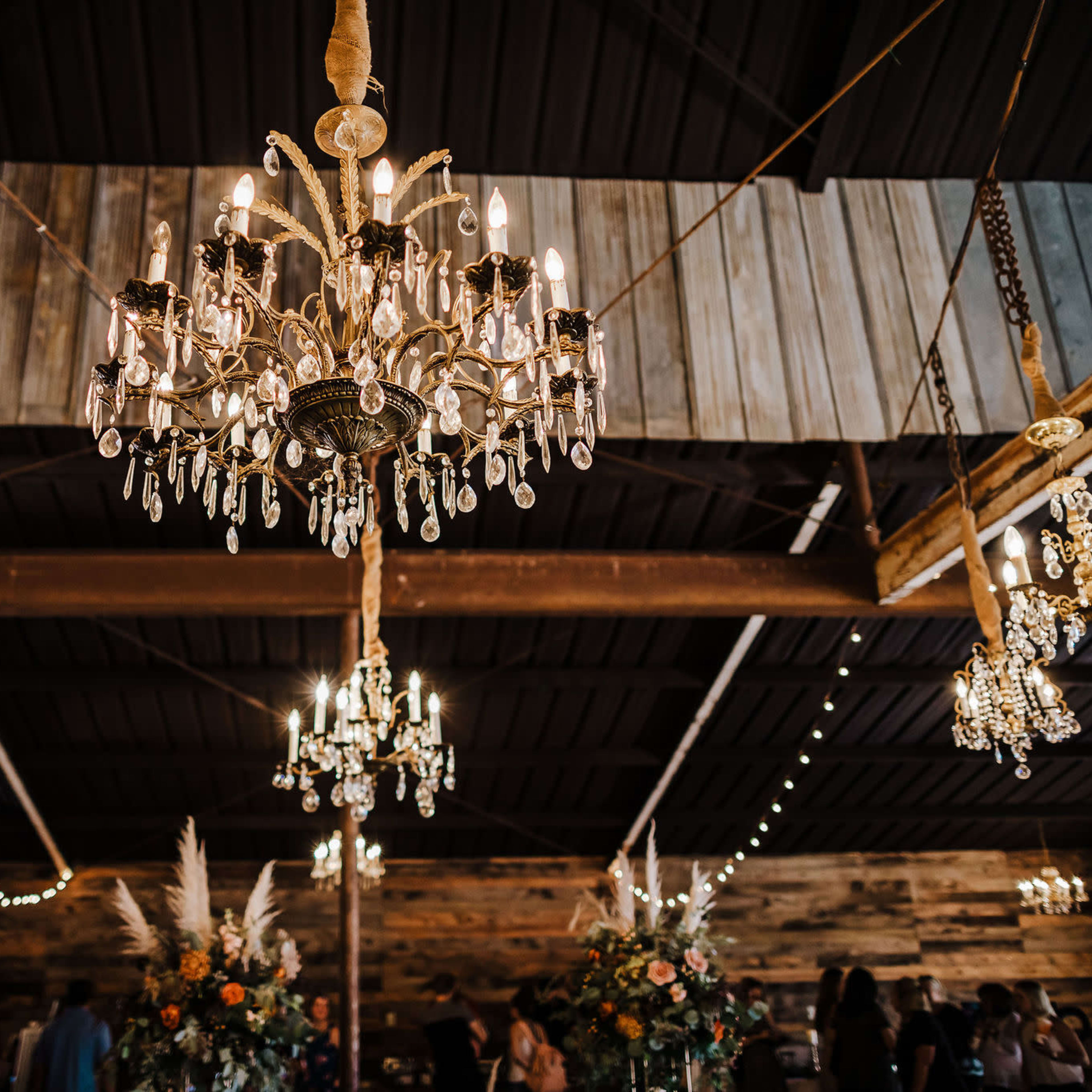 A rustic interior with multiple ornate chandeliers hanging from a wooden ceiling, adorned with string lights and decorated tables.