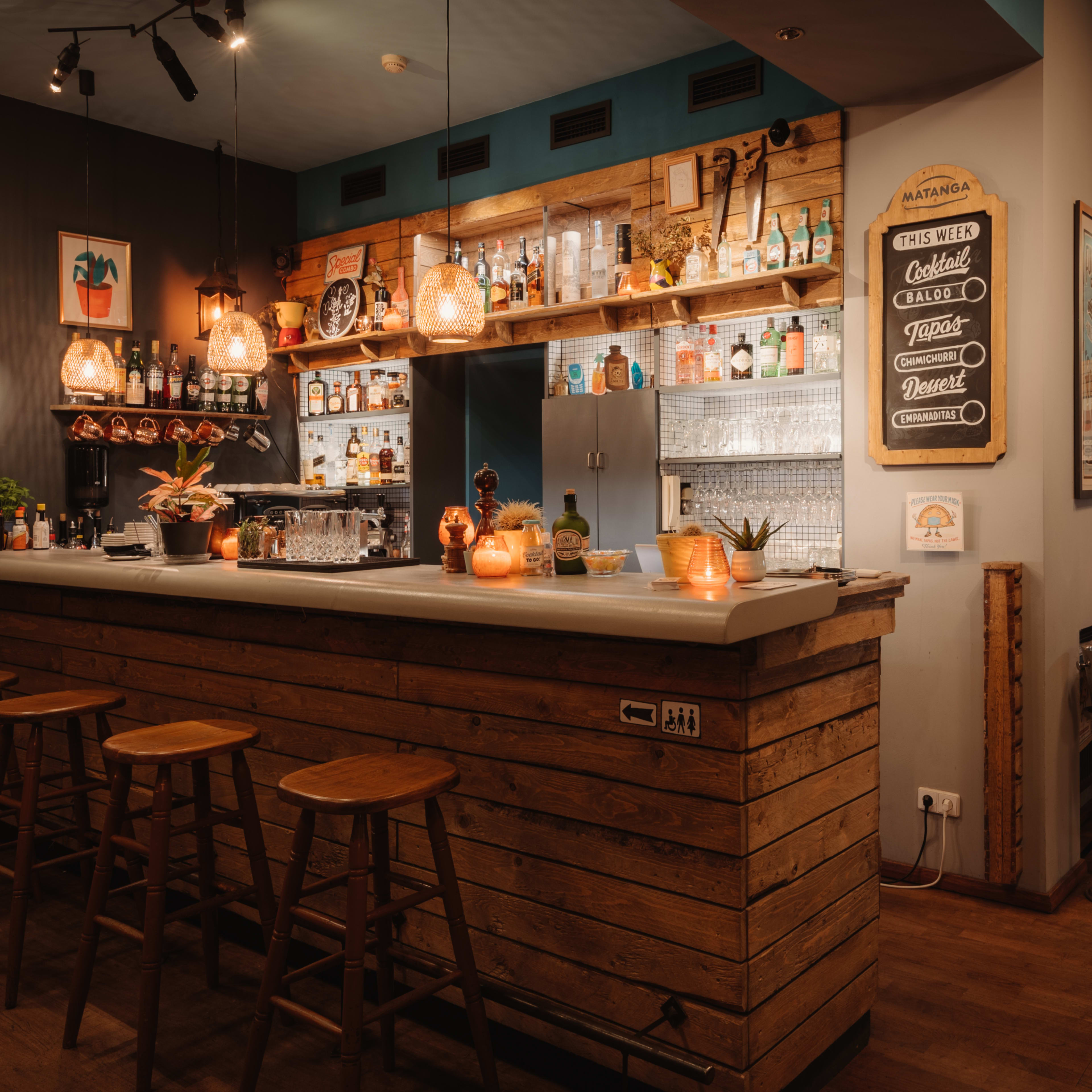 A rustic bar area with wooden stools, a variety of bottles on shelves, and softly glowing candles on the counter.