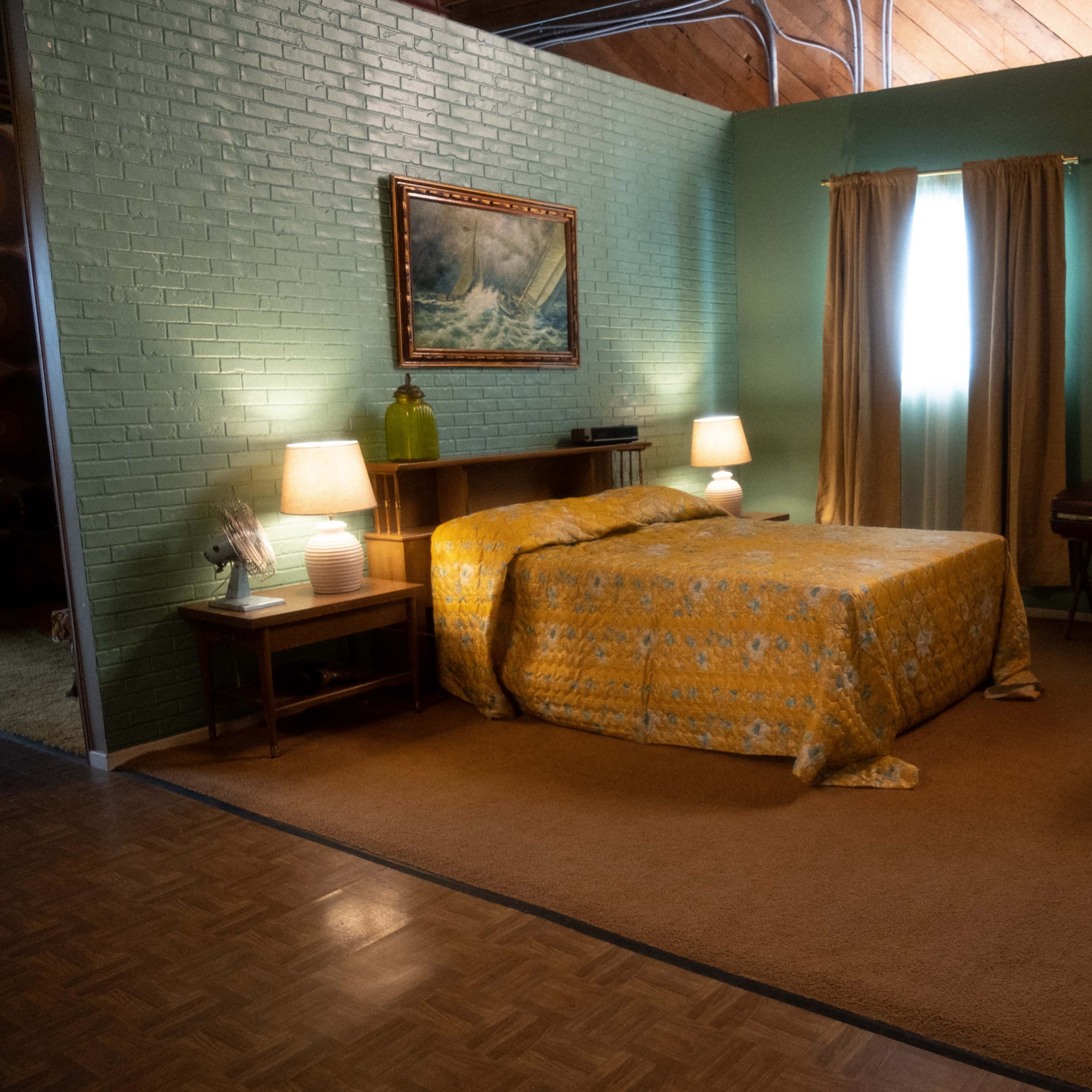 A bedroom with a bed covered in a yellow patterned blanket, a wooden desk, and a door leading to another room, all set against a green brick wall.