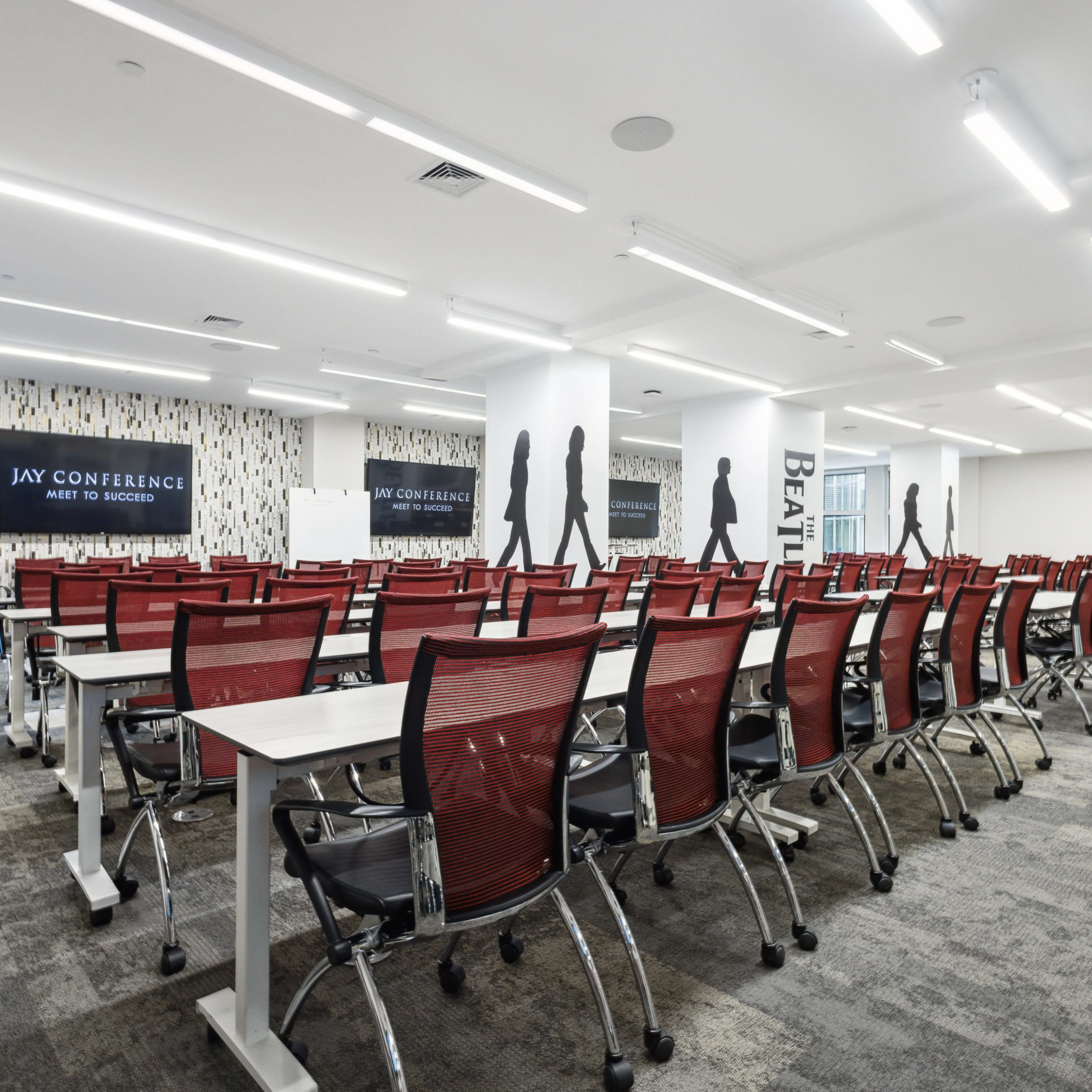 A modern conference room features rows of red seated tables arranged for a meeting, with wall graphics and ample overhead lighting.