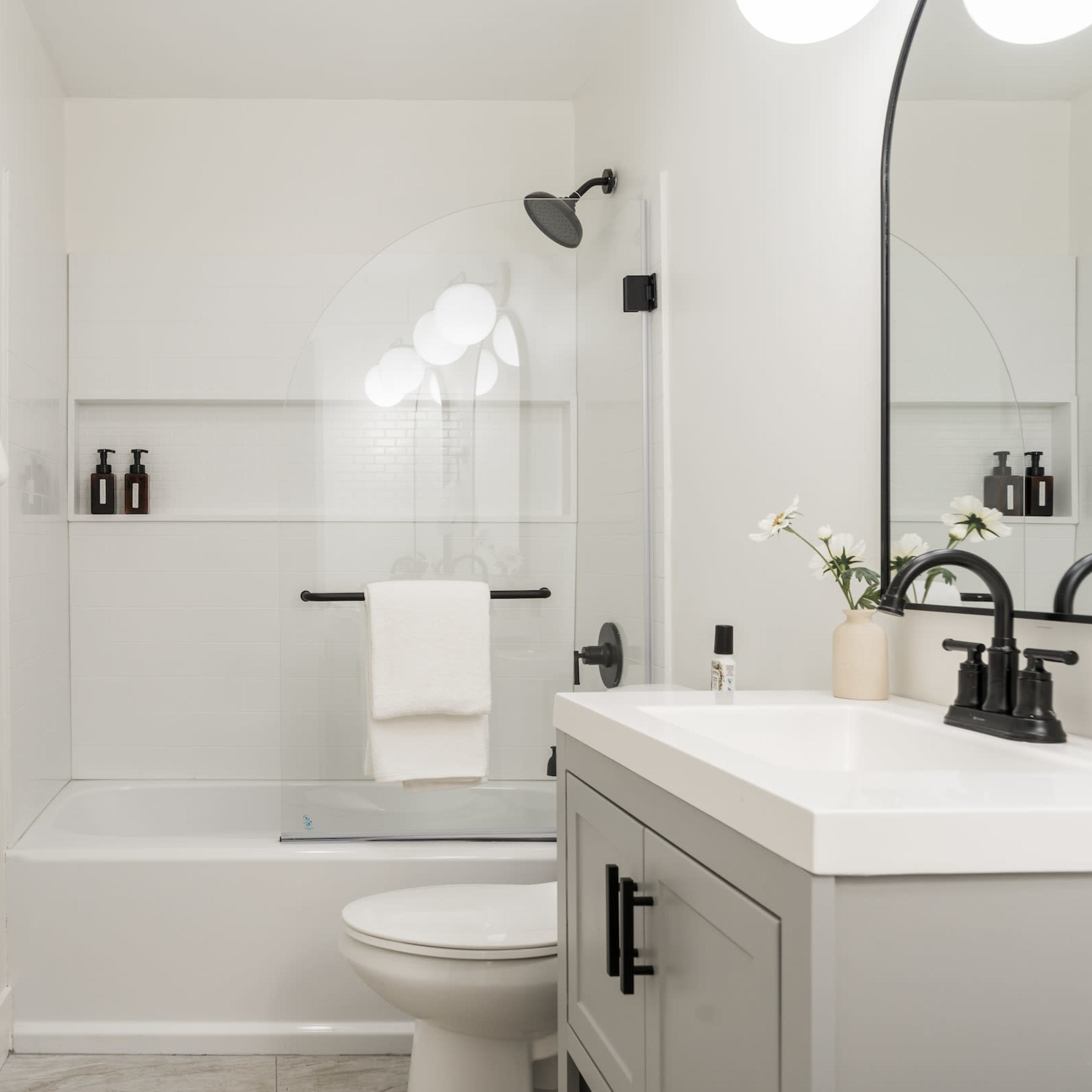 A modern bathroom featuring a glass shower enclosure, a white countertop vanity with black fixtures, and light-colored walls.