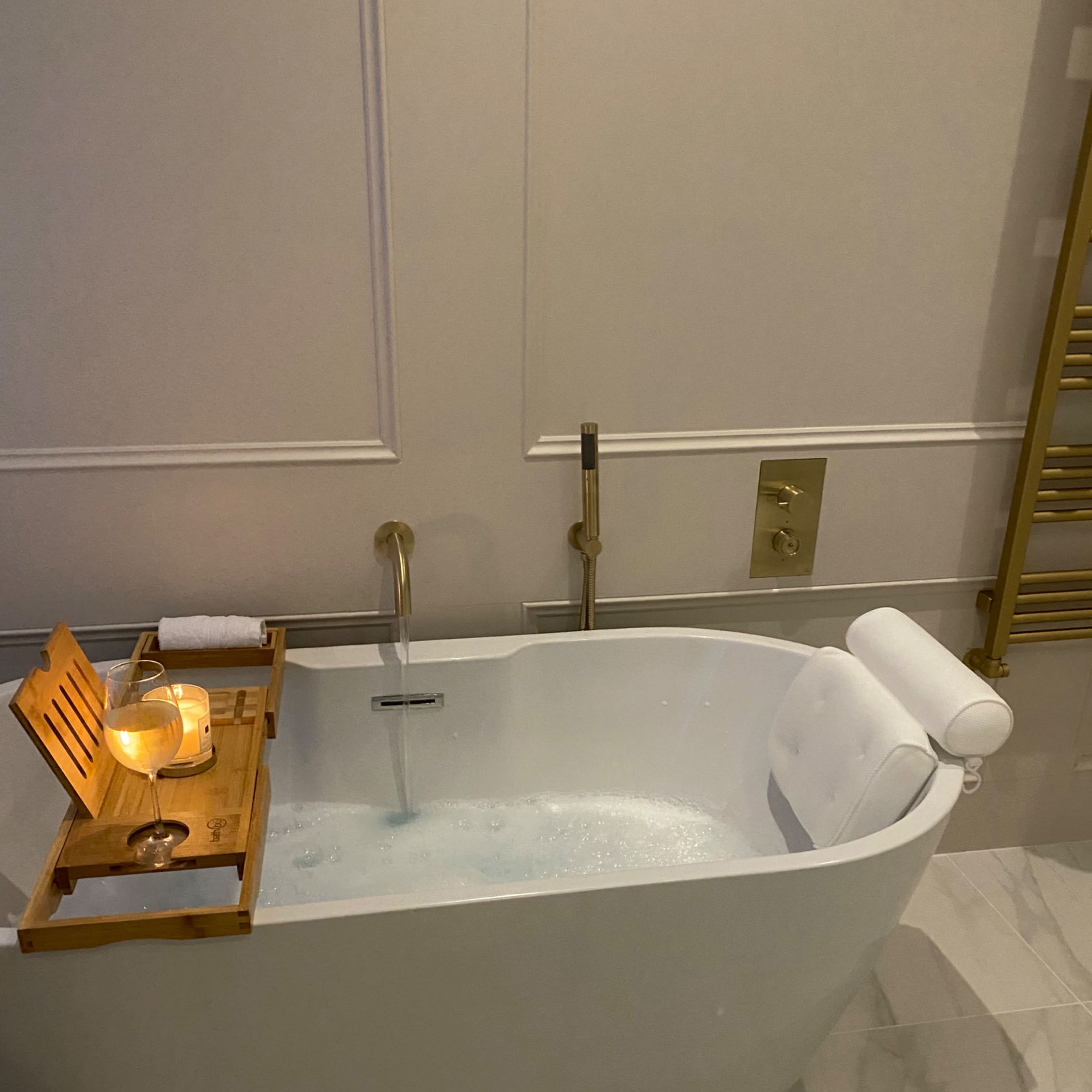 A modern white freestanding bathtub with a wooden tray holding a candle, placed in a room with marble flooring and a gold towel rack.