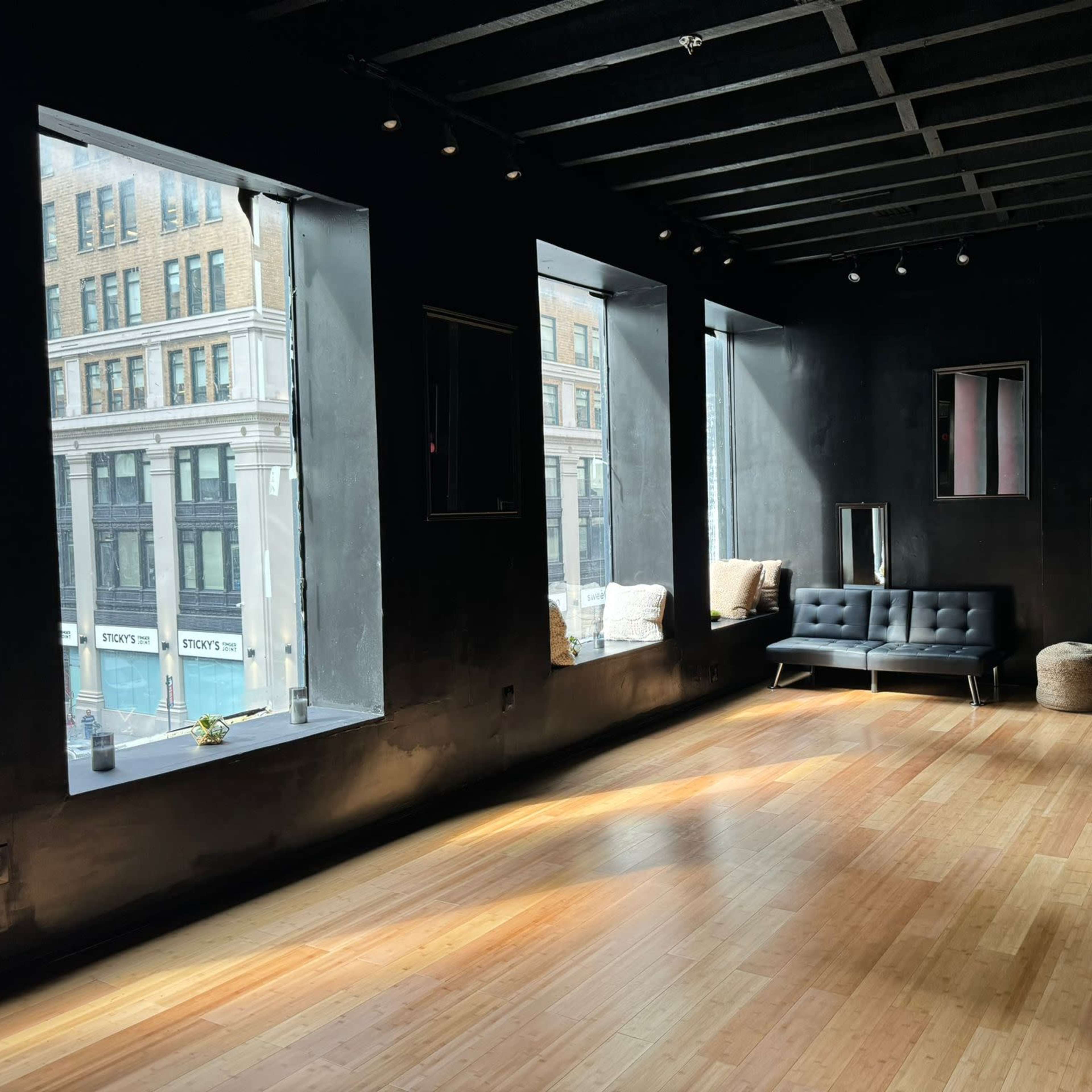 The room features large windows that allow natural light to illuminate a space with dark walls and wooden flooring, furnished with black couches and a few seating areas.