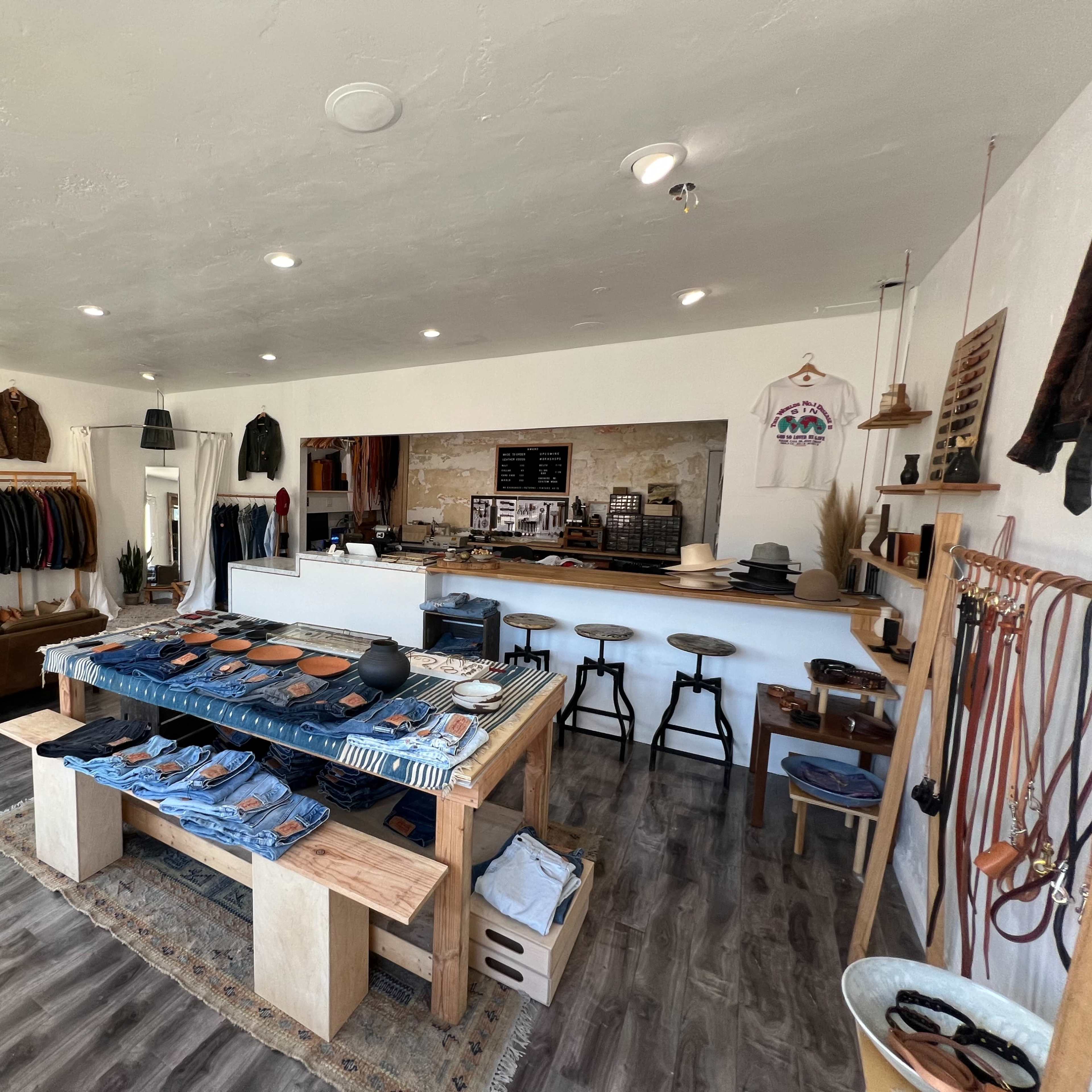 An interior of a clothing store featuring racks of apparel, a central display table with denim items, and a minimalist wooden counter area.
