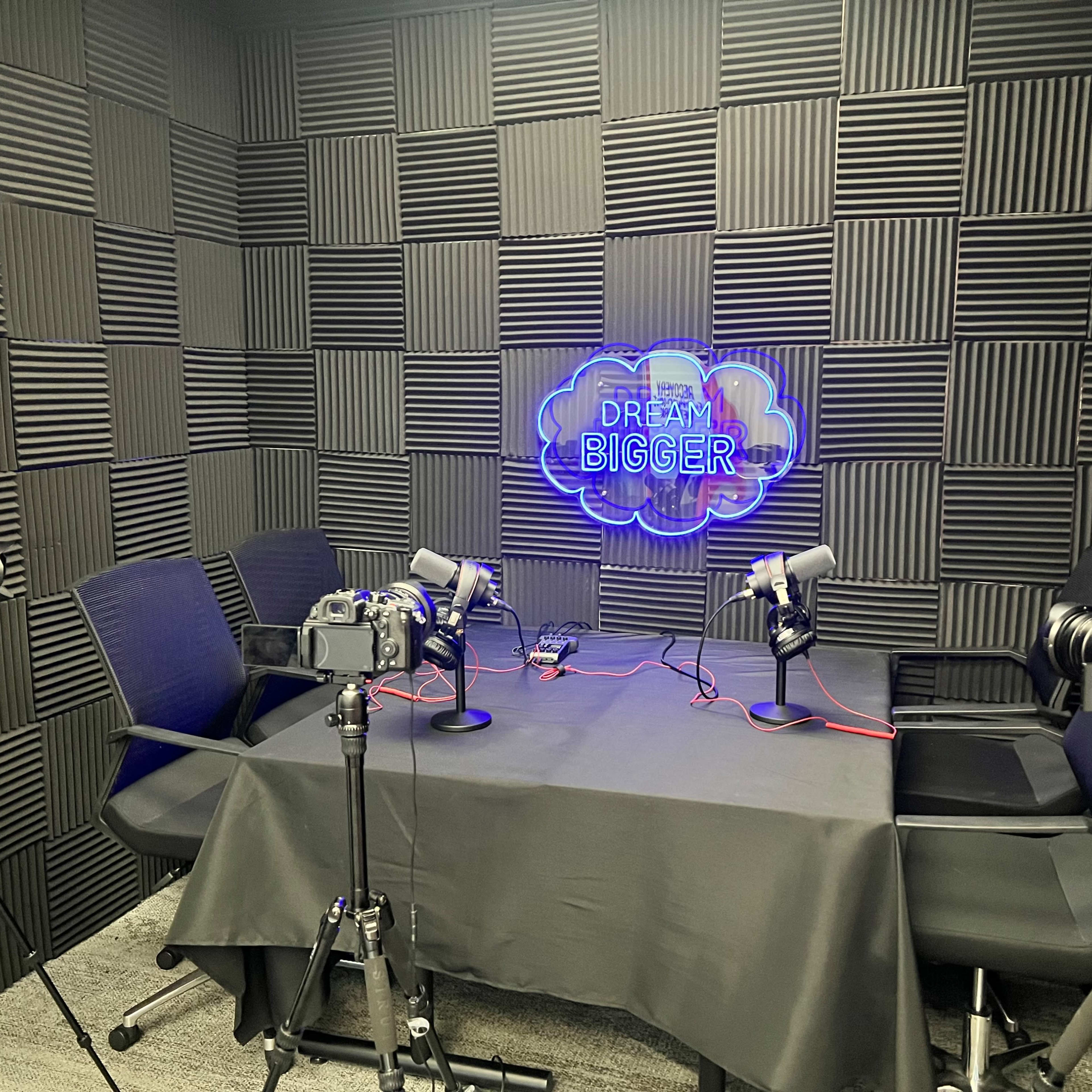 A podcasting studio featuring two microphones, two cameras, and a neon sign on a textured wall.