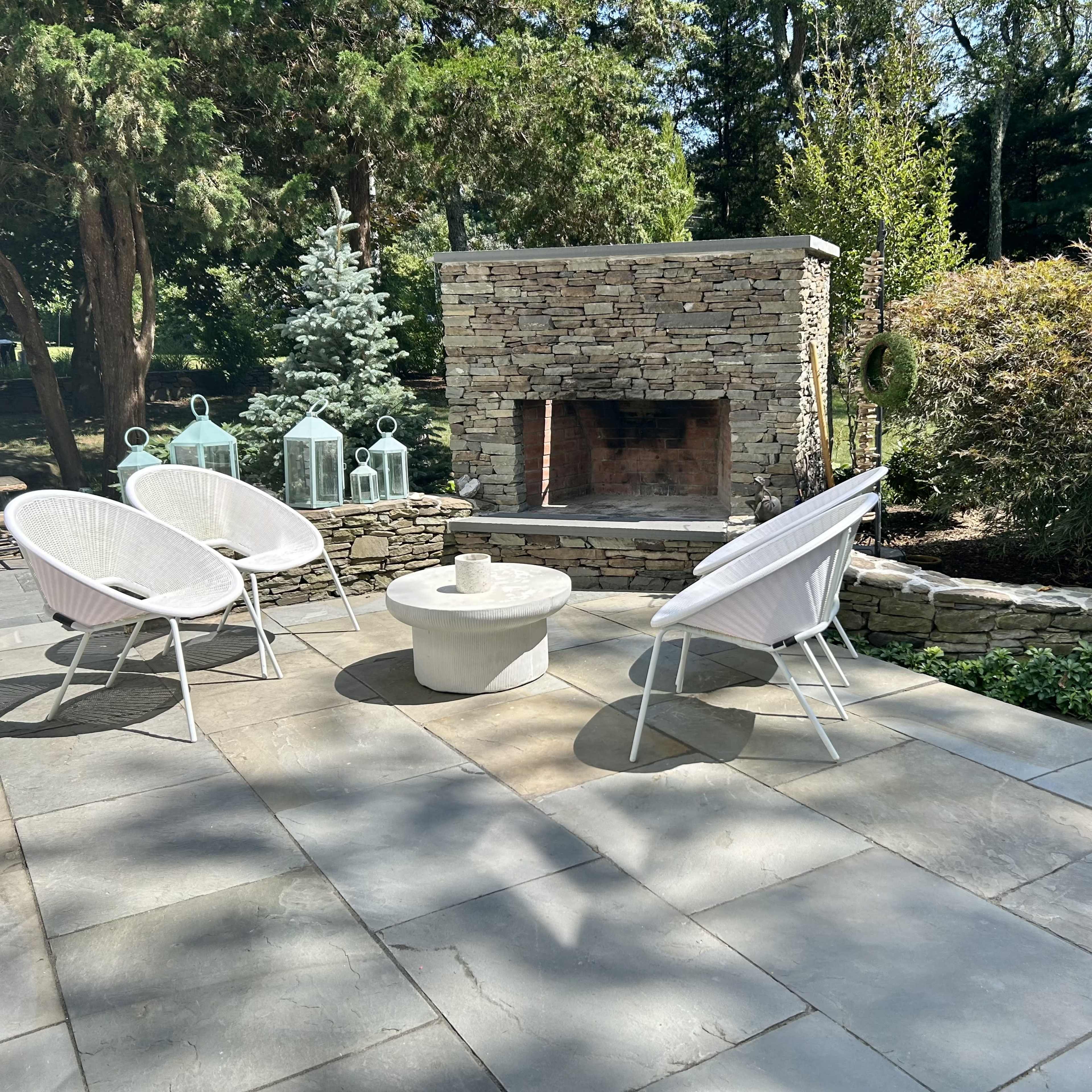 A stone patio area with three whitechairs arranged around a circular table, complemented by a stone fireplace and lush greenery in the background.