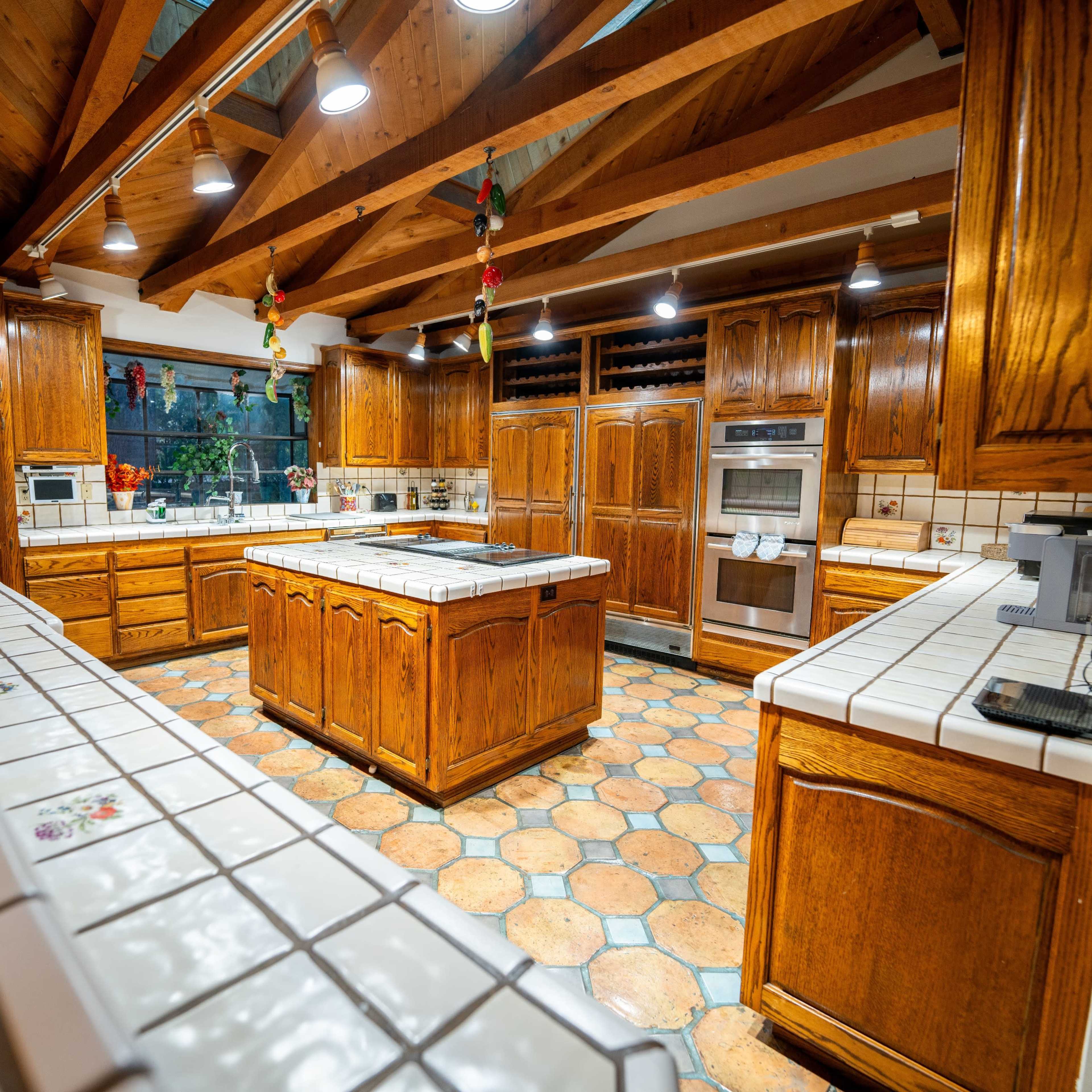 A spacious kitchen with wooden cabinets, tile countertops, and a central island, illuminated by overhead lights.