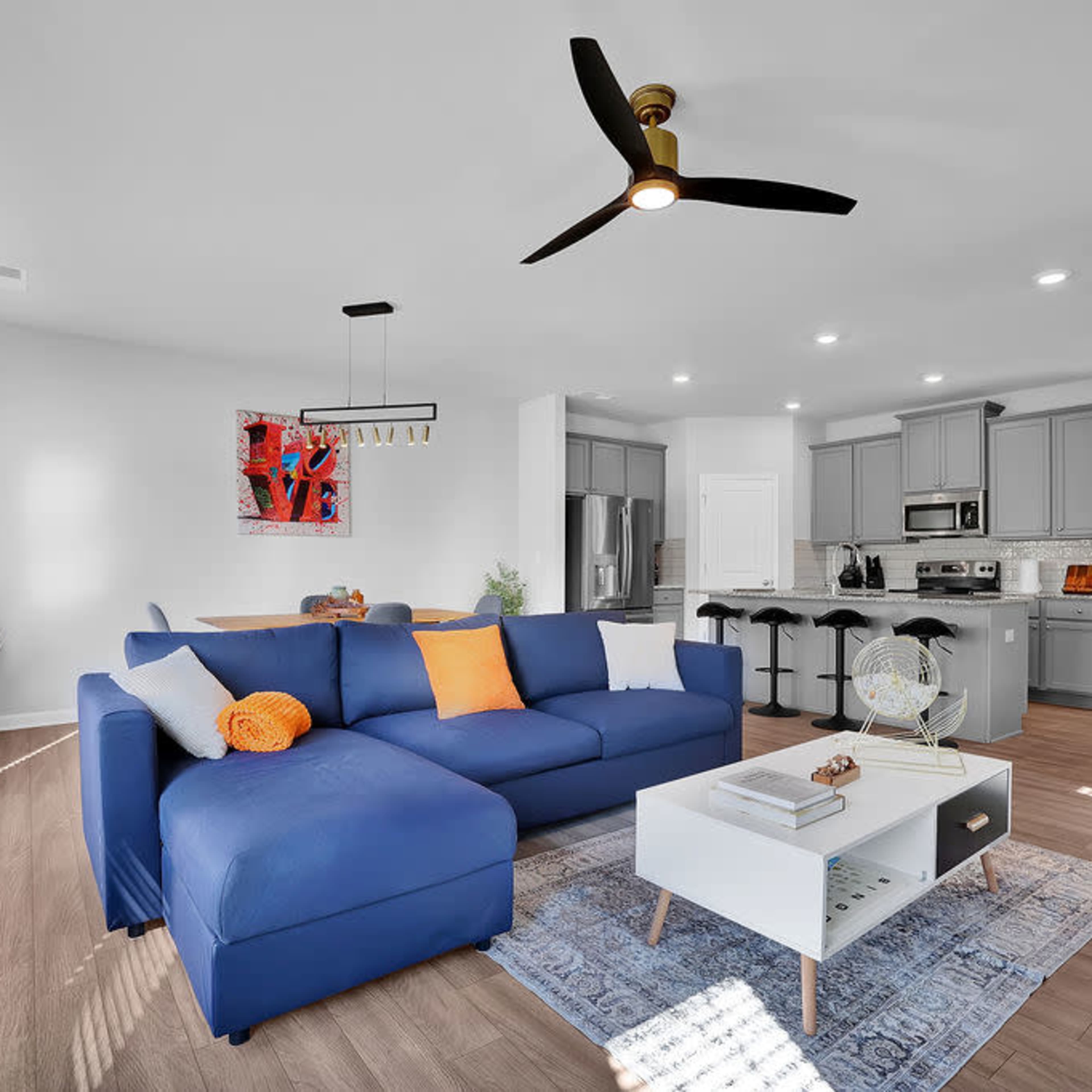 A spacious living room features a blue sectional sofa, a coffee table, and an open kitchen with gray cabinets and stainless steel appliances.