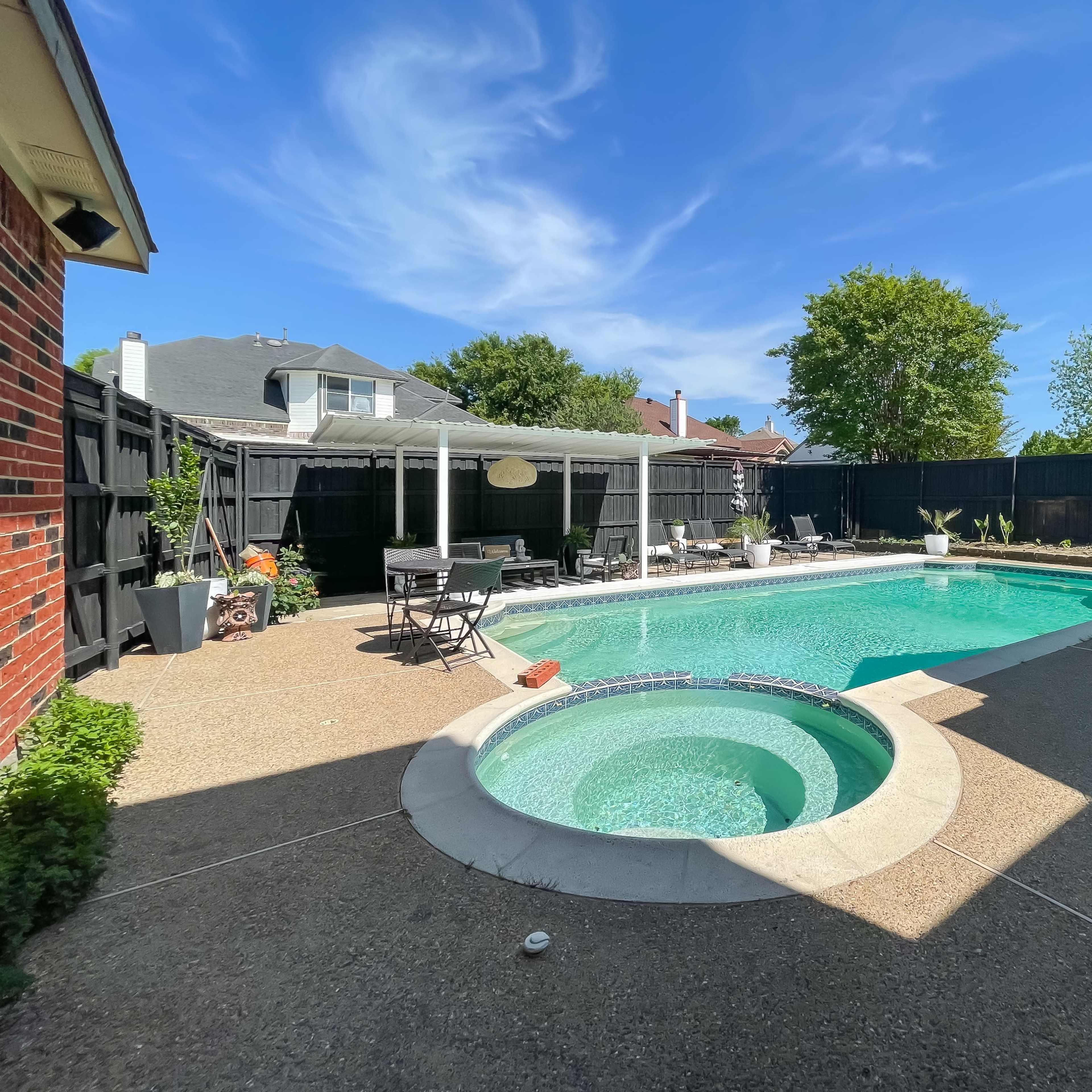 A backyard swimming pool with a built-in hot tub, surrounded by a patio area and greenery under a clear blue sky.