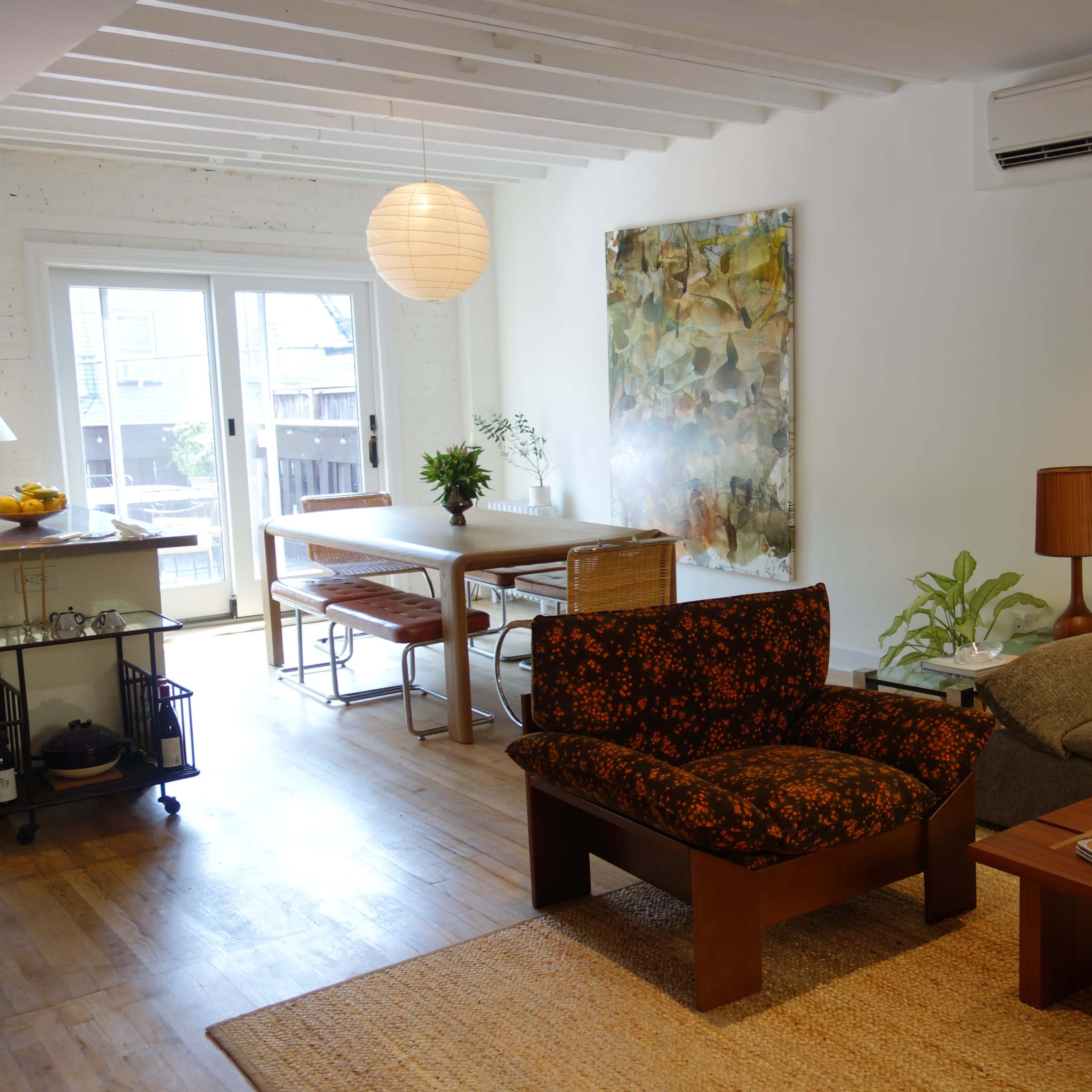 A modern living space with a brown floral-patterned armchair, a gray sofa, a wooden coffee table, a dining area with a table and chairs, and large windows leading to an outdoor space.