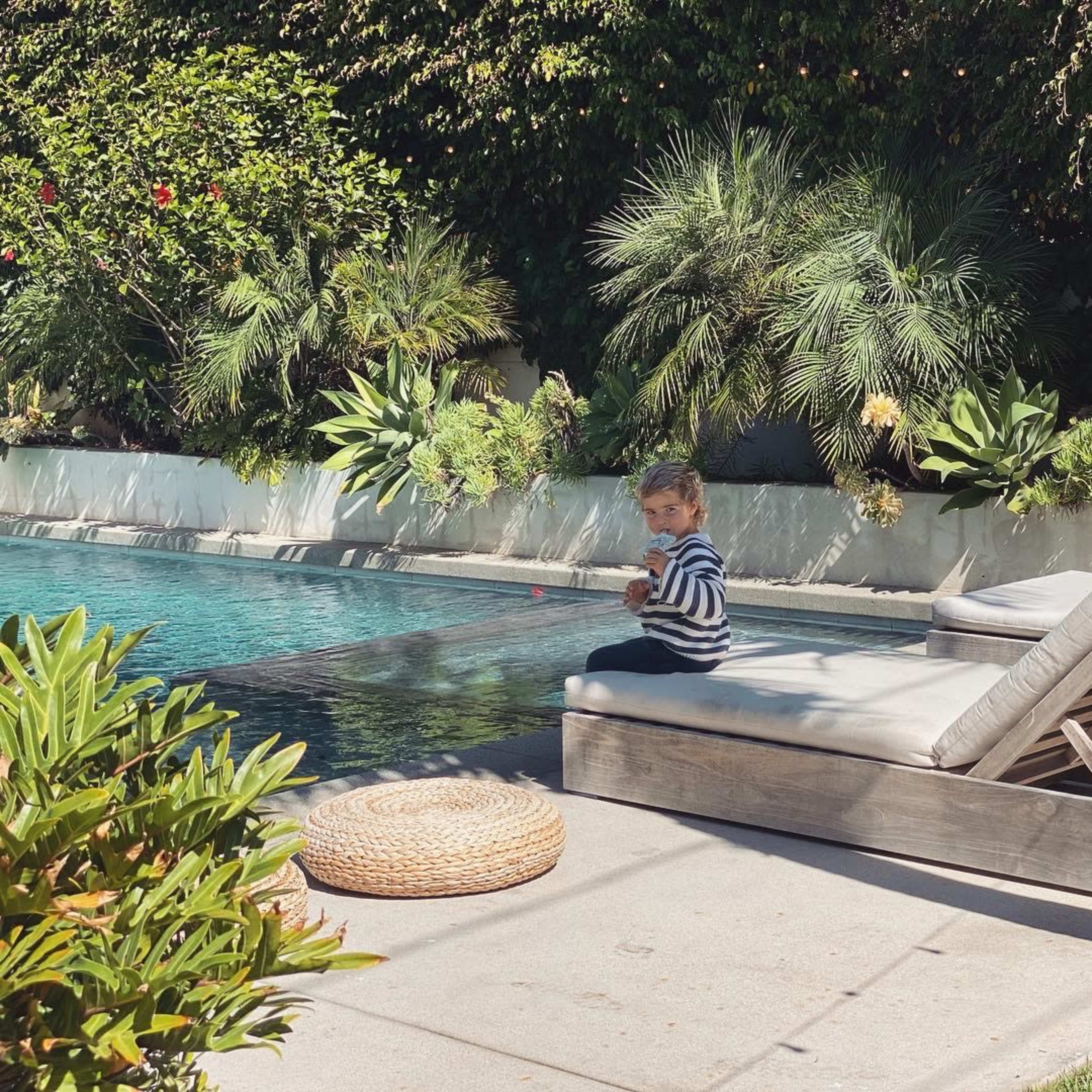 A child in a striped shirt sits on the edge of a sunbed beside a swimming pool surrounded by green plants.