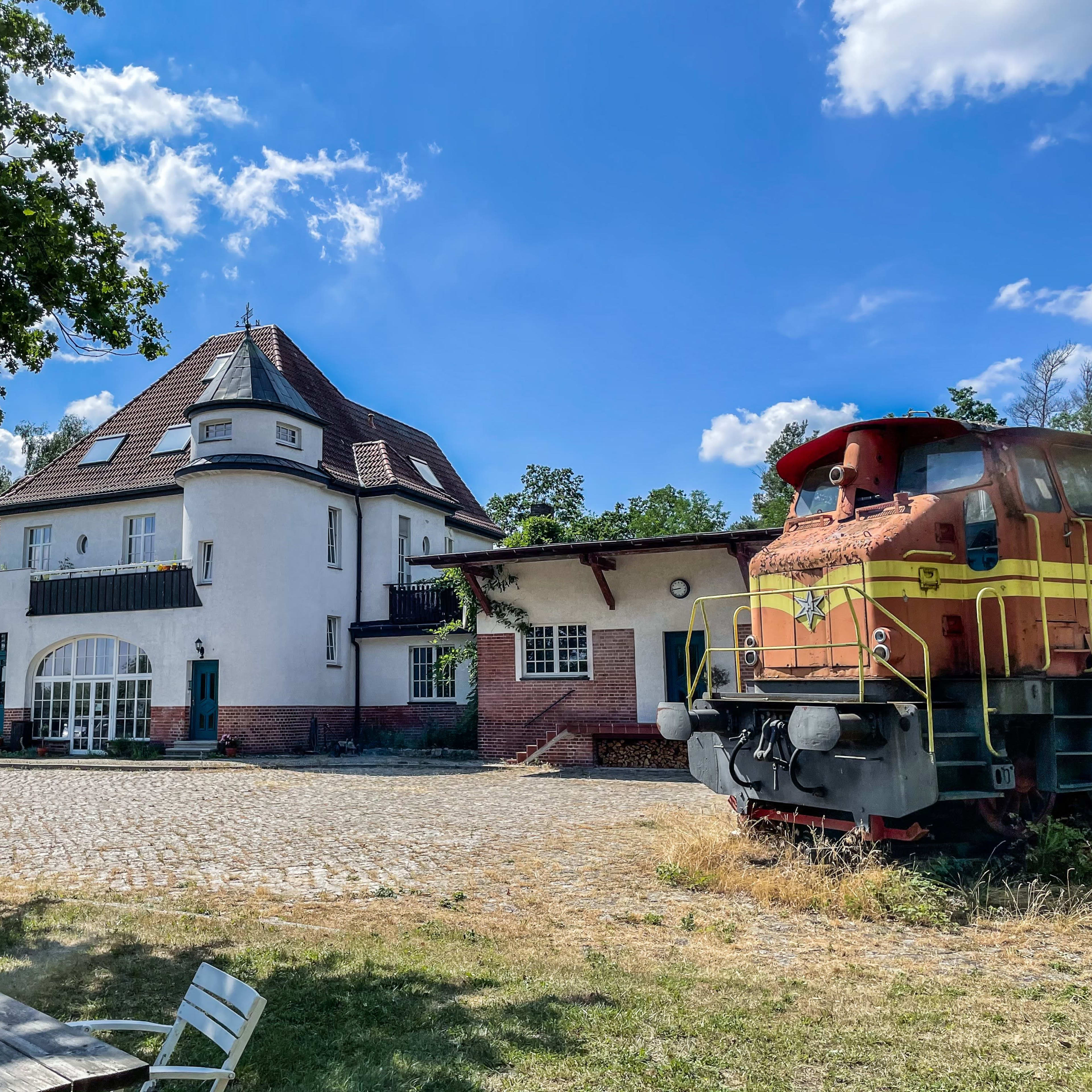 A large, white, three-story building adjacent to a parked train locomotive on a gravel area under a blue sky with scattered clouds.