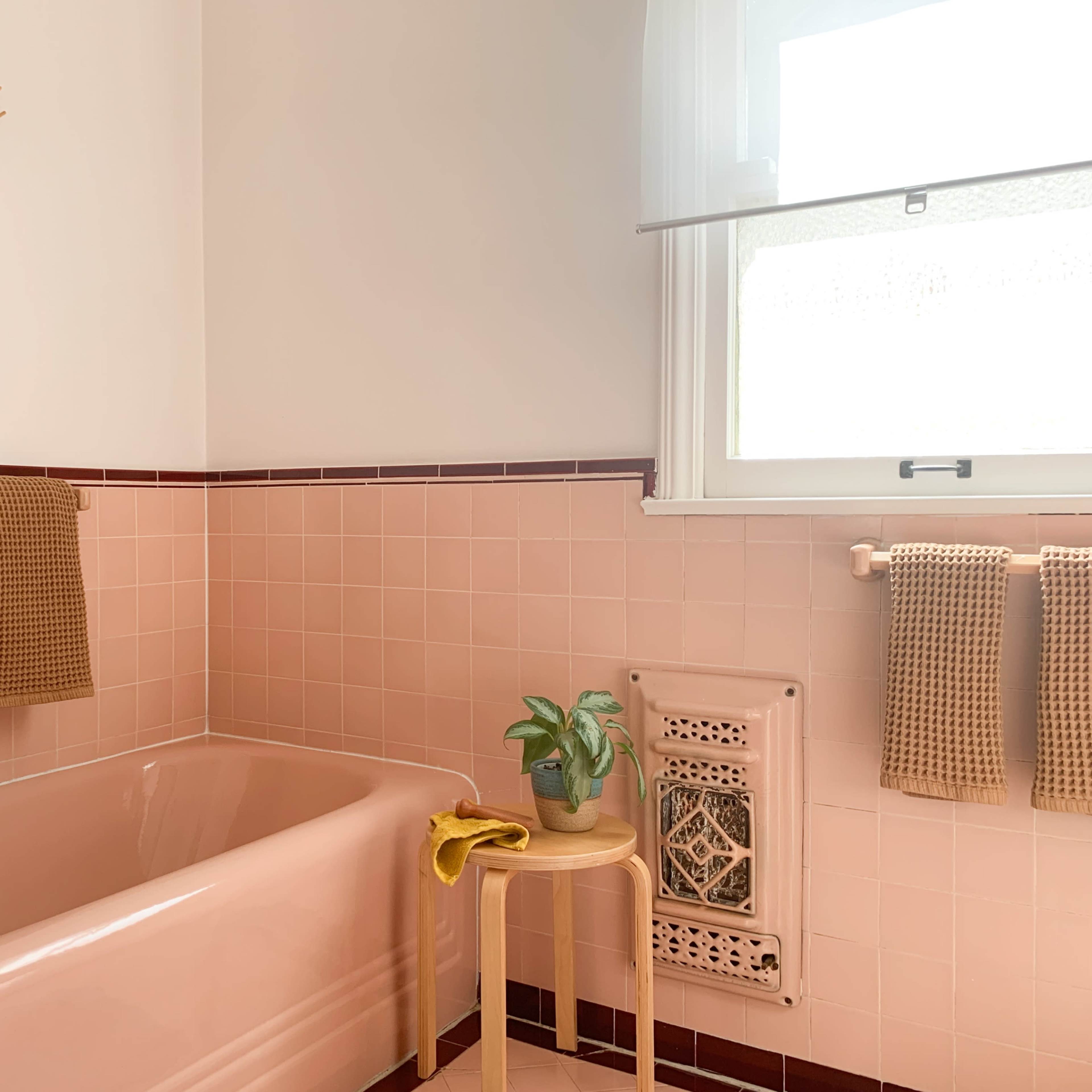 A bathroom featuring a pink bathtub, tiled walls, and brown towels hanging beside a small plant on a metal table.