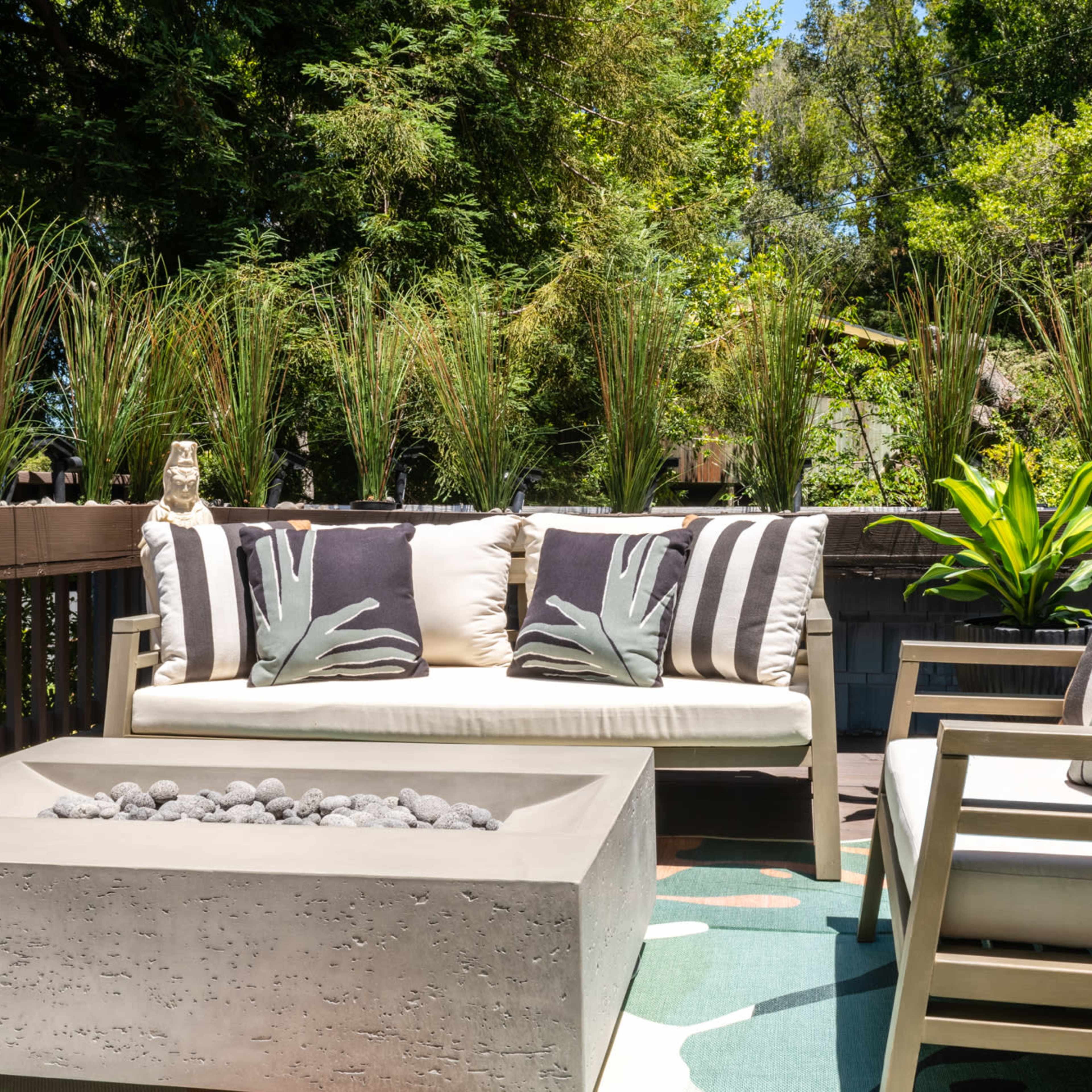 A patio area with a light-colored sofa and chairs, featuring decorative pillows and a fire pit table surrounded by tall greenery.