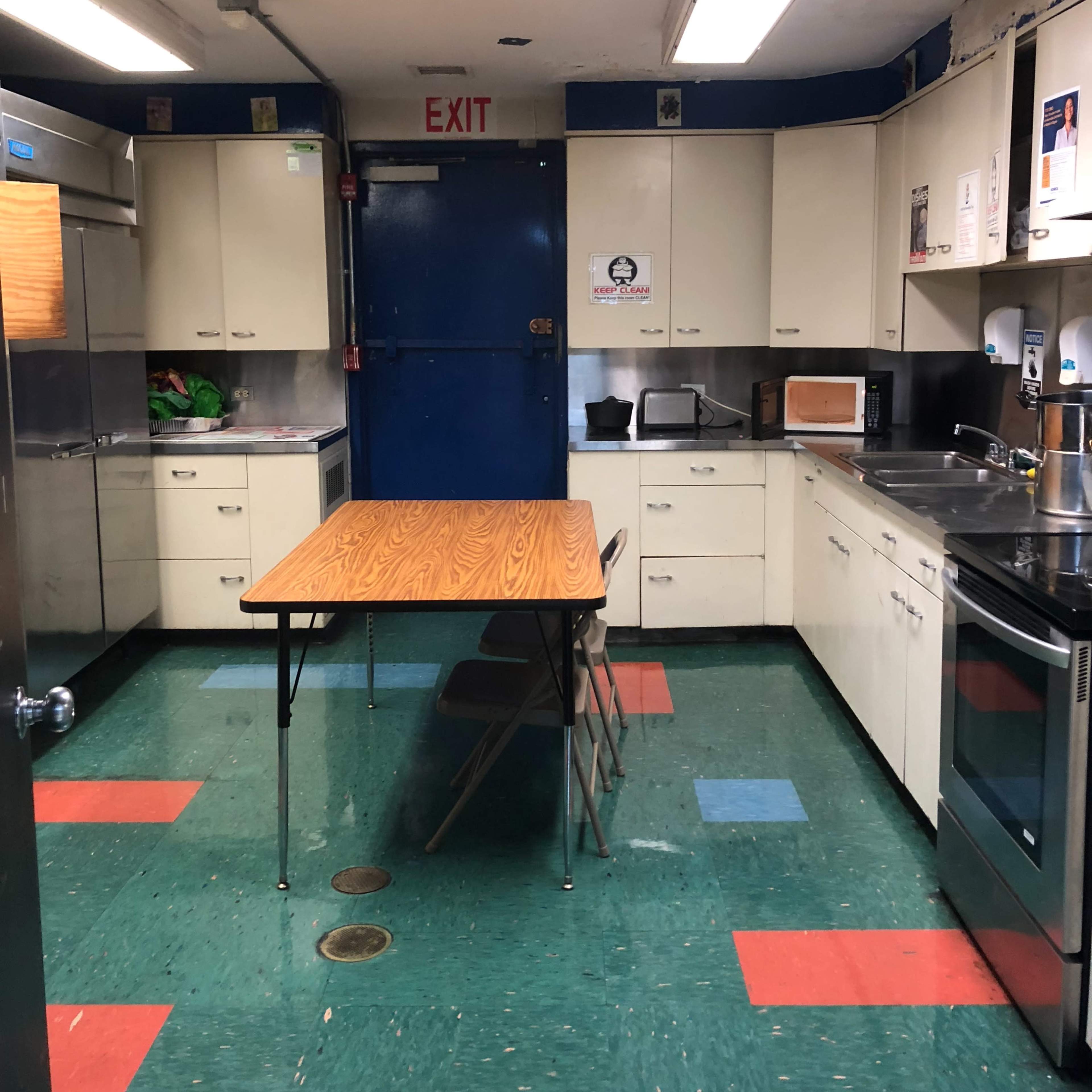 A kitchen with a central table and two chairs, surrounded by cabinets, appliances, and an exit door.