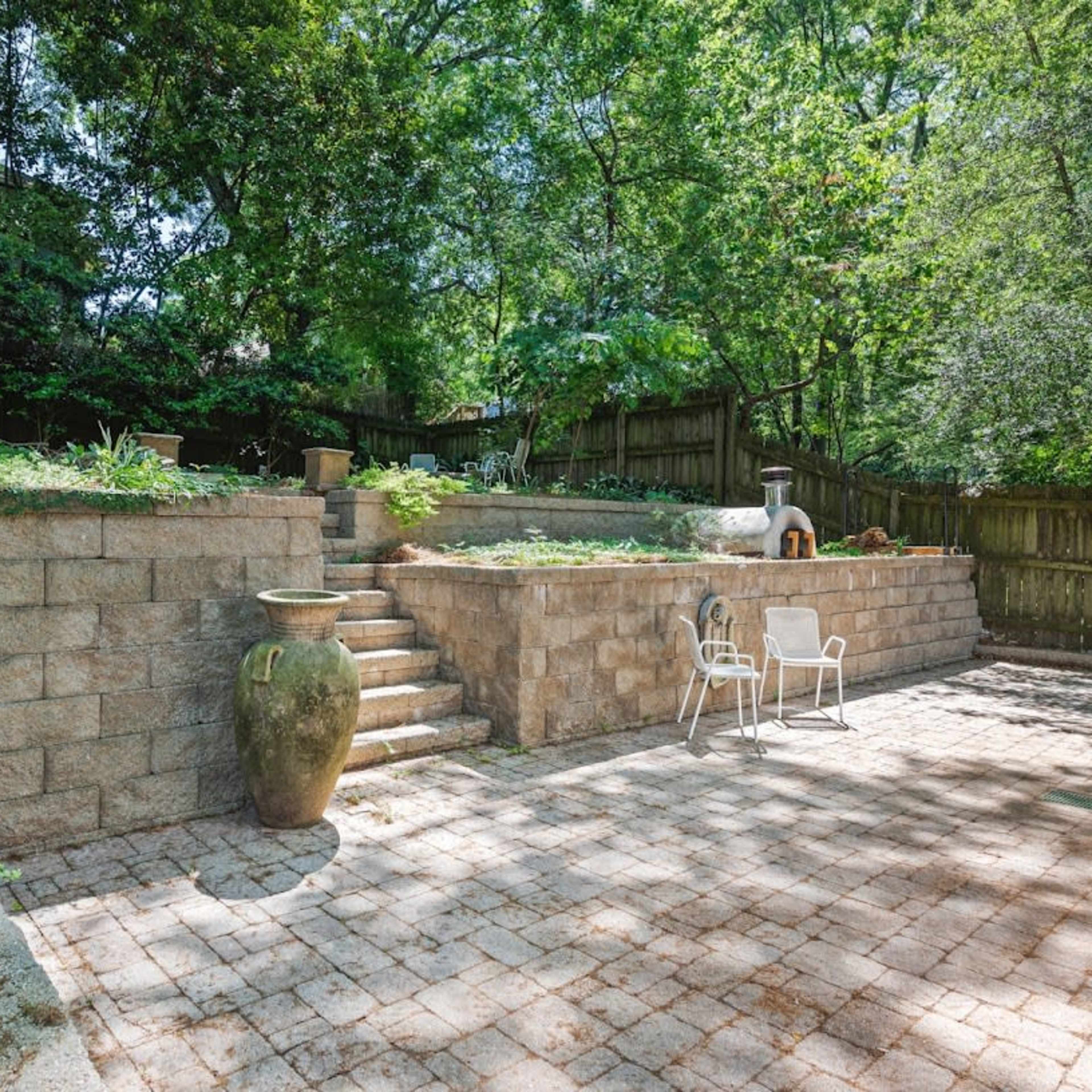 A stone patio features steps leading to a raised garden area surrounded by greenery and a wooden fence.