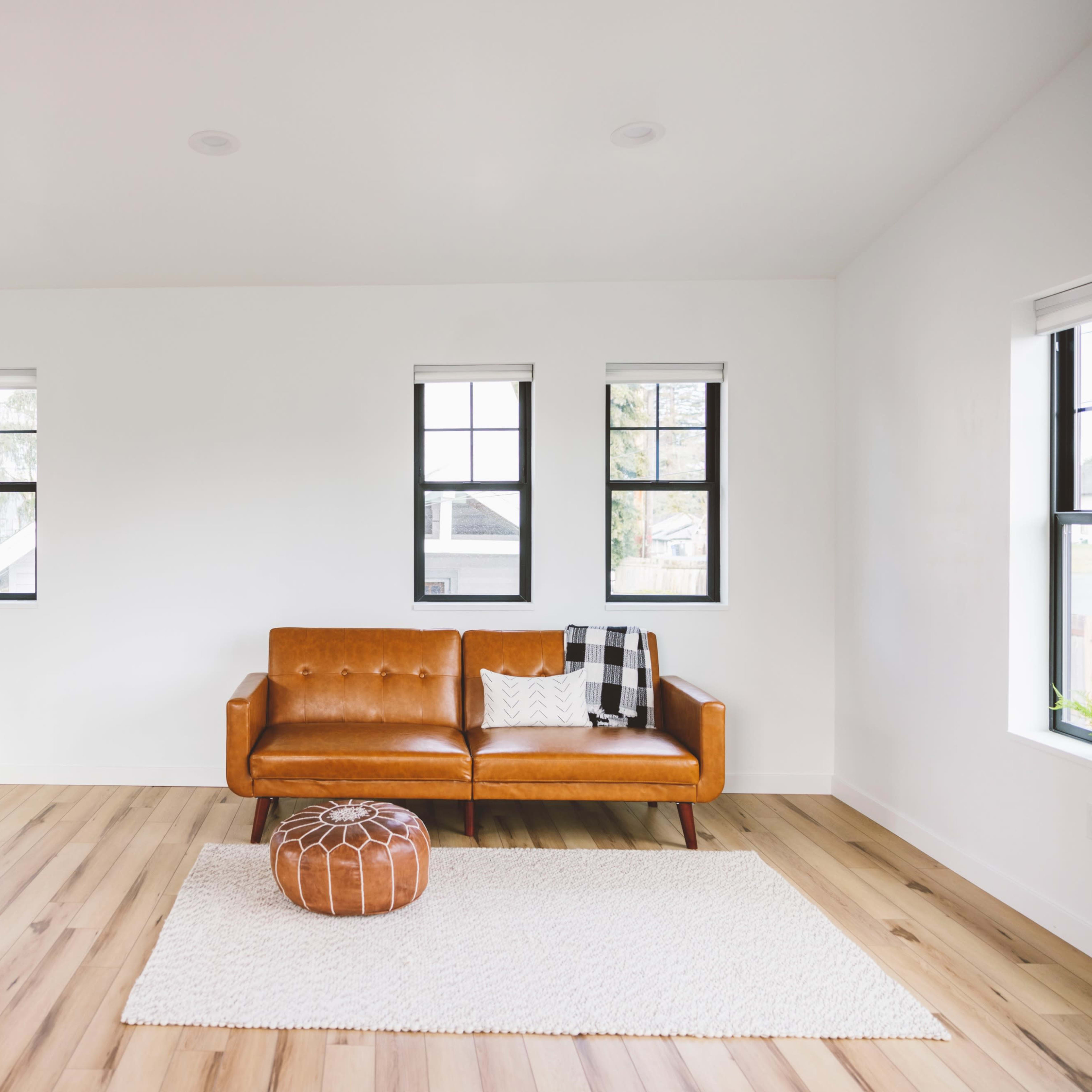 A modern living room features a brown leather sofa, a round pouf, and a light area rug surrounded by large windows and a potted plant.