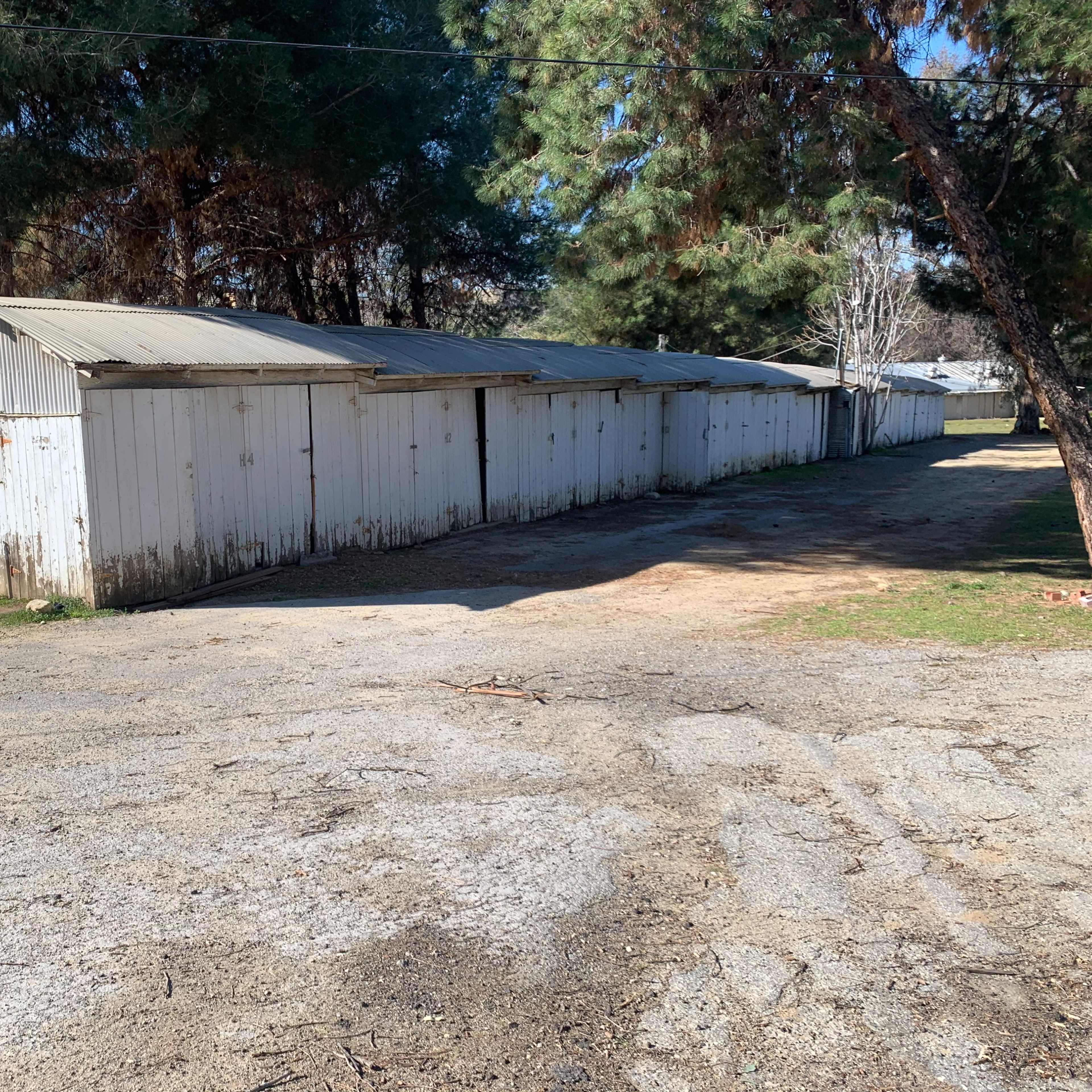 A row of weathered white storage sheds situated along a gravel path in a wooded area.