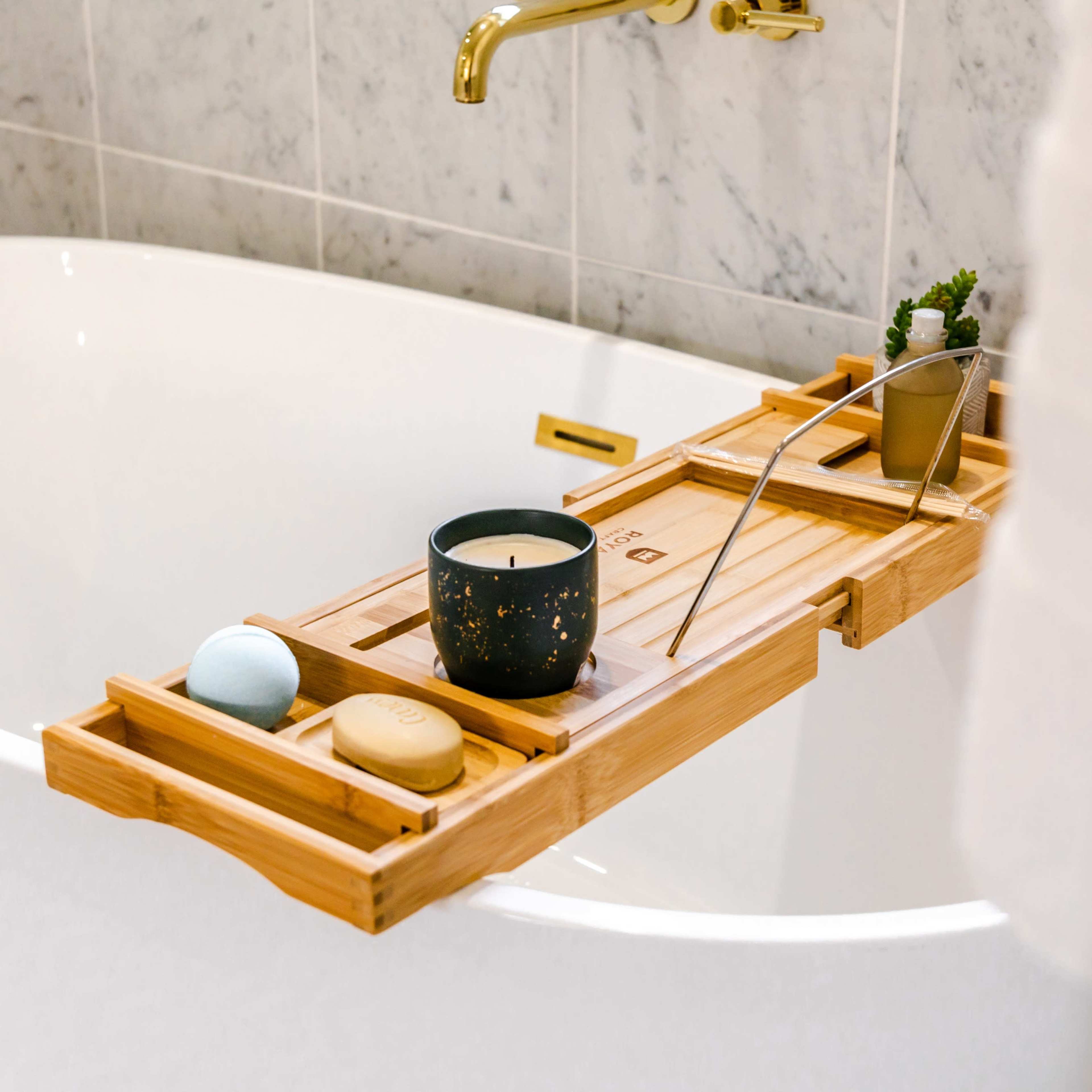 A wooden bath tray is placed across the edge of a white bathtub, holding a candle, a small potted plant, and several bath products.