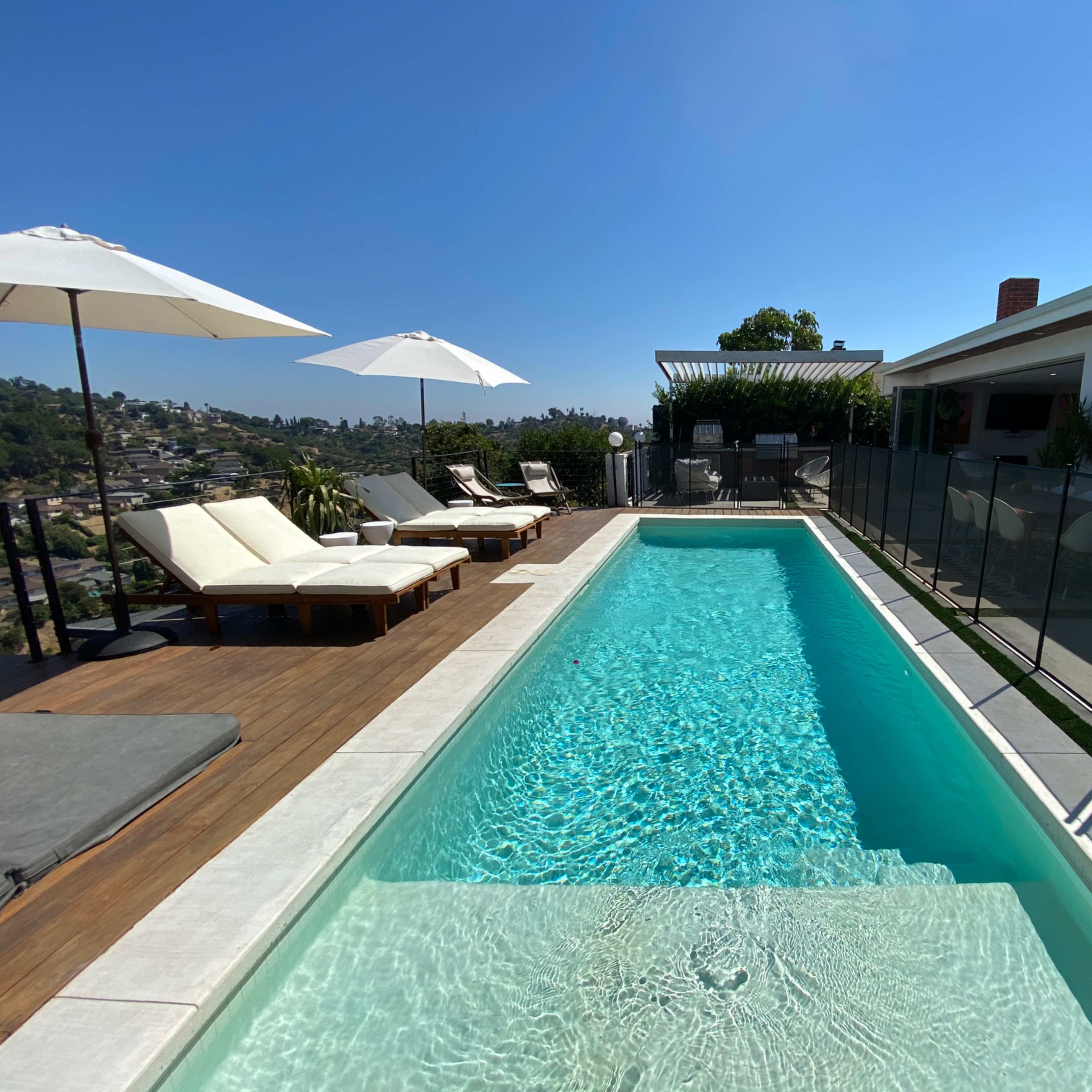 A modern outdoor living space with white furnishings, a pool in the background, and a dog near a landscaped area overlooking a scenic hillside.