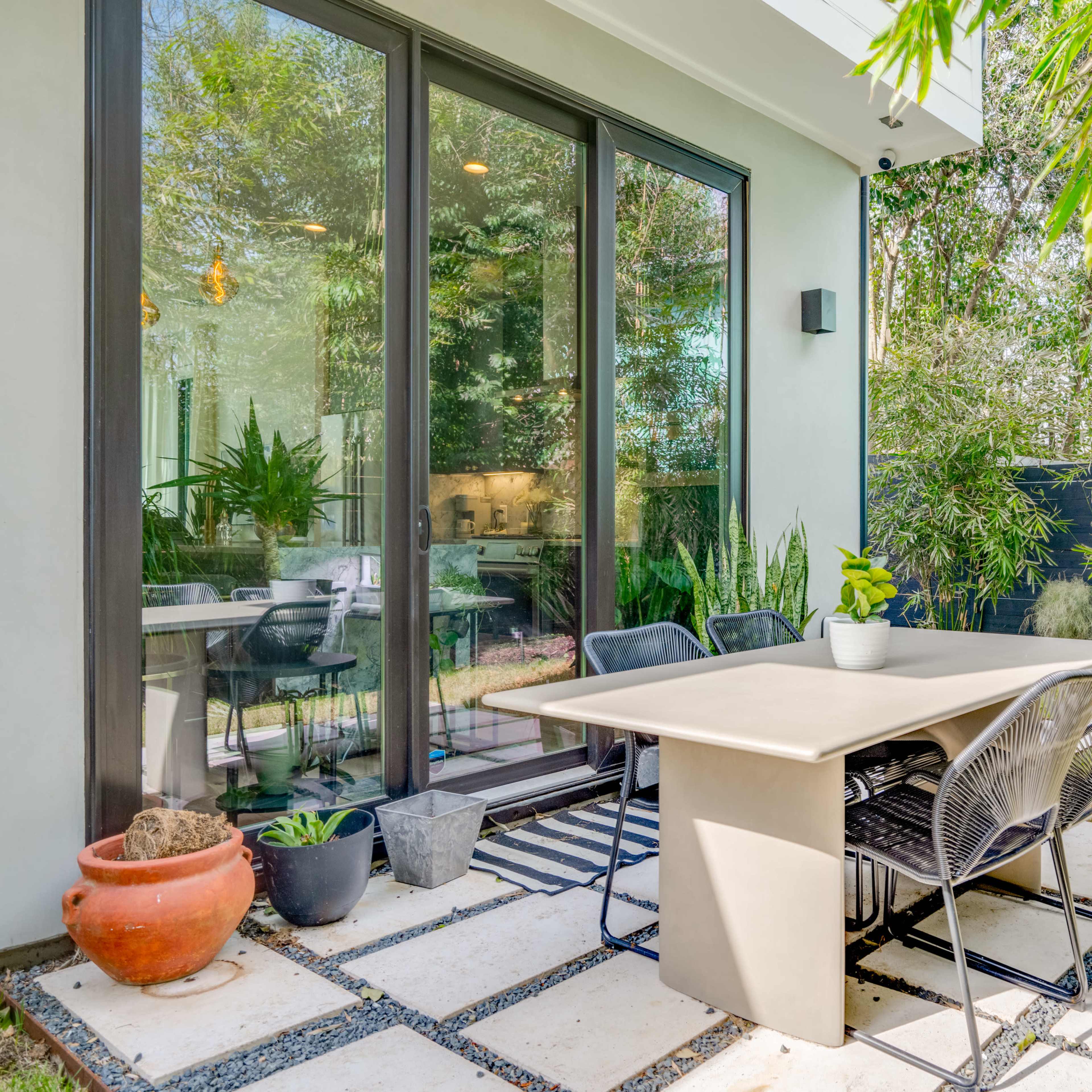 A patio area features a table and chairs surrounded by greenery and a view of large windows.
