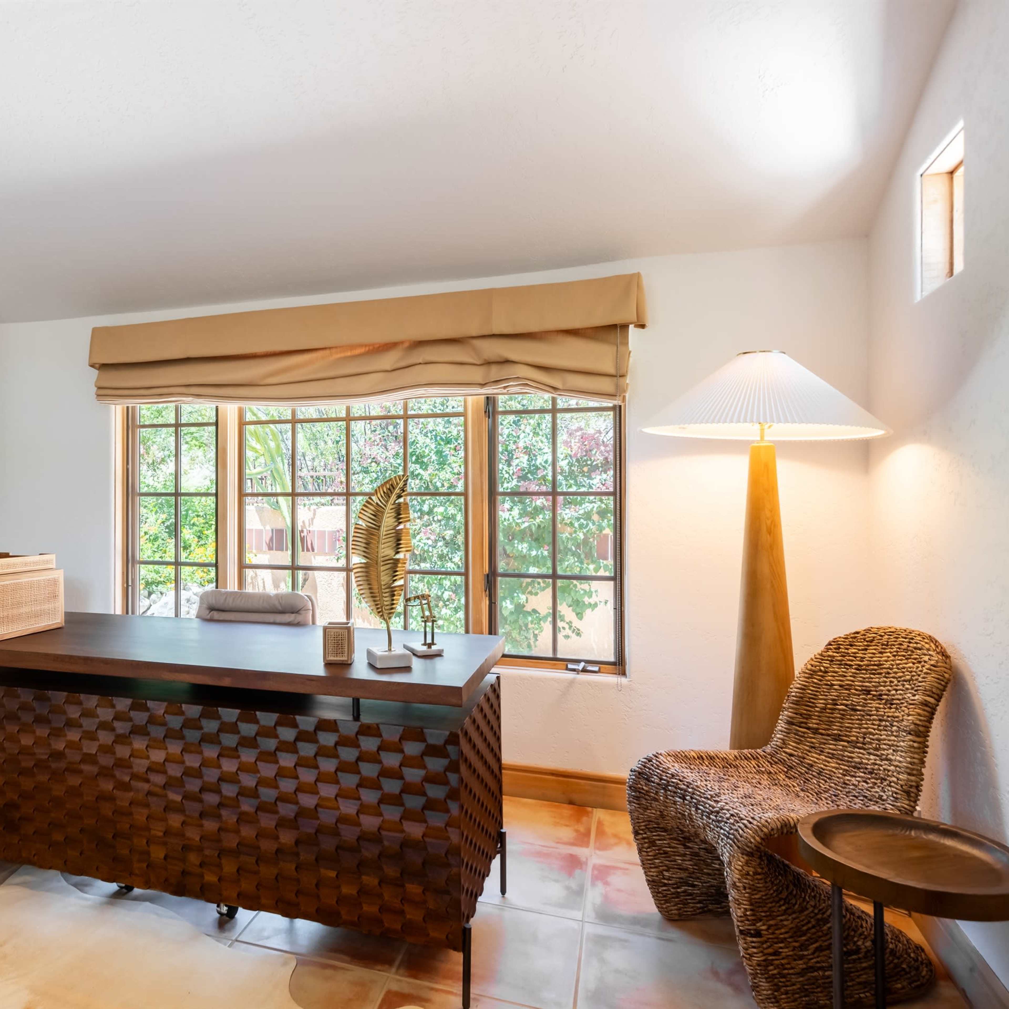 A well-lit home office with a wooden desk, a wicker chair, a standing lamp, and large windows overlooking greenery.