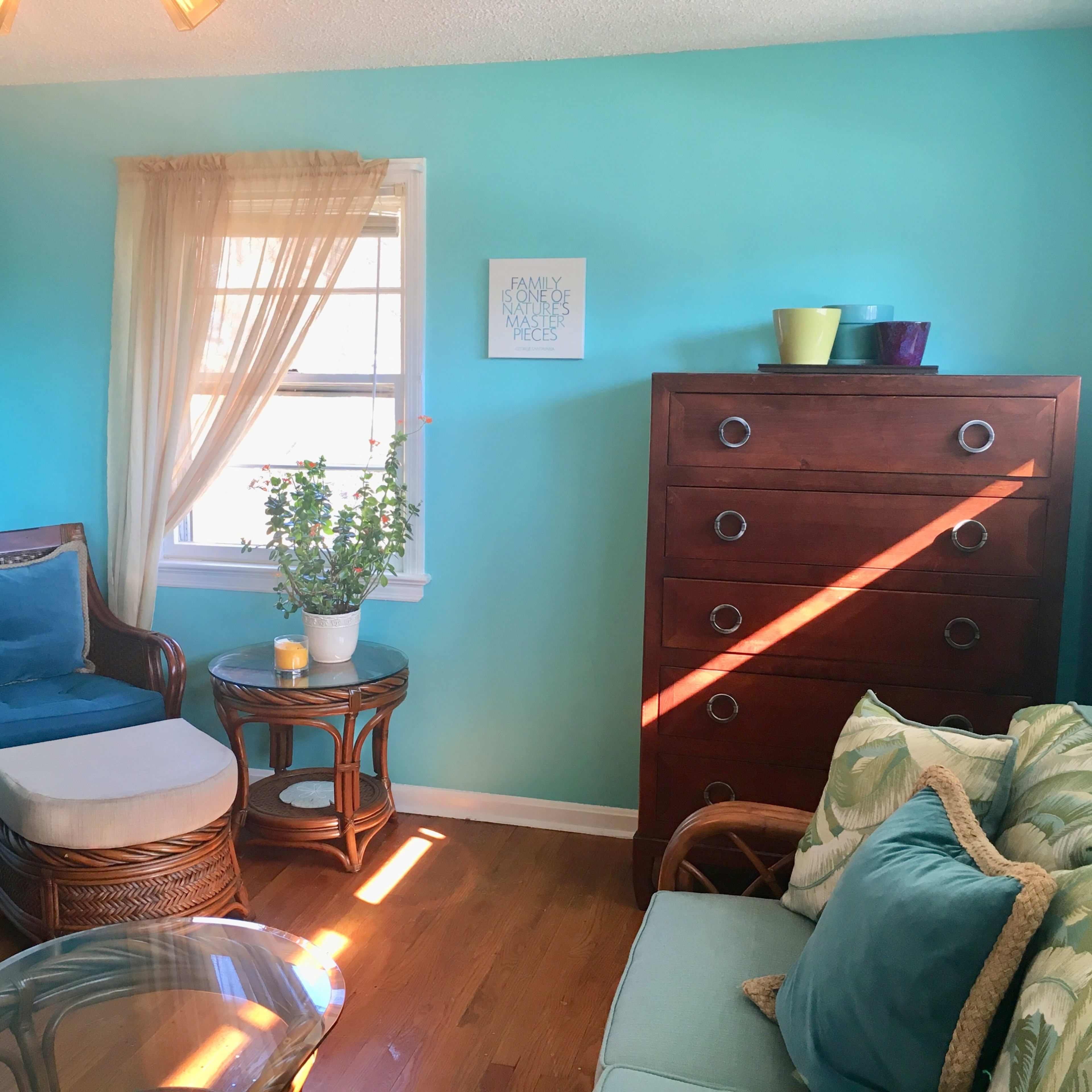 A bright room with light blue walls, a wooden dresser, a plant on a table, and various colorful decorative items on top of the dresser.