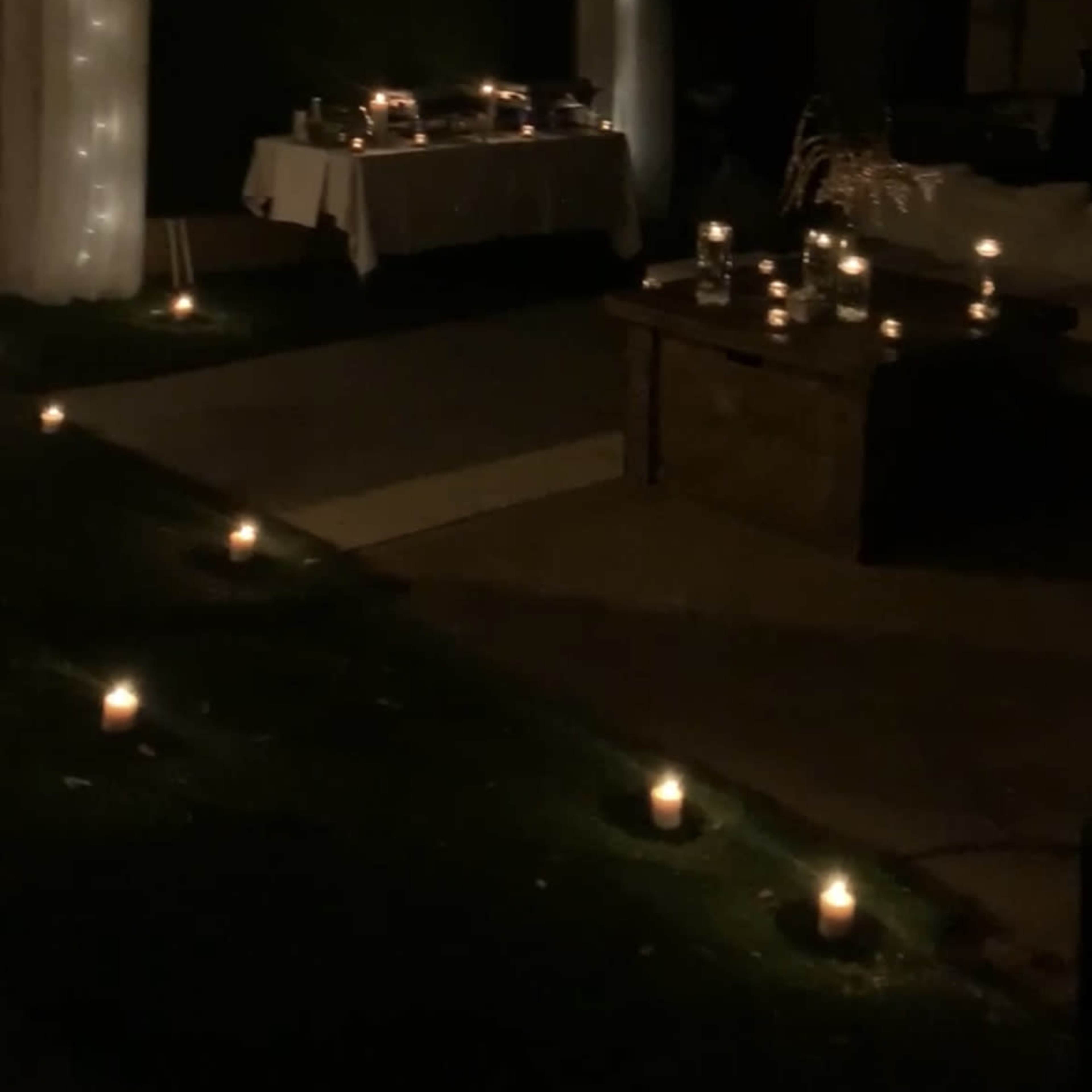 The scene features a dimly lit outdoor area with tables set for an event, surrounded by candles on the ground.