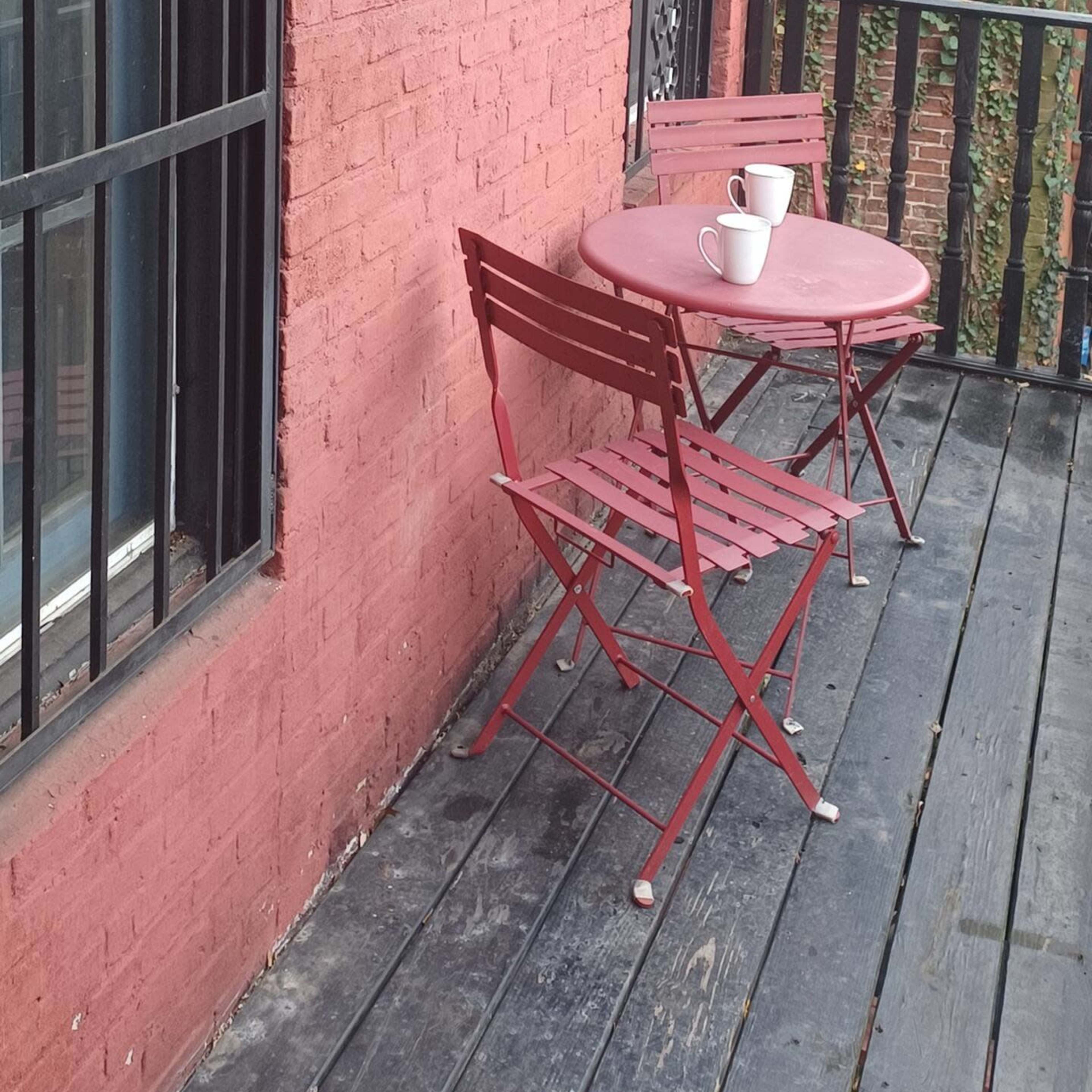 A red brick wall features a small red table and two chairs on a wooden balcony alongside a window with decorative metal bars.