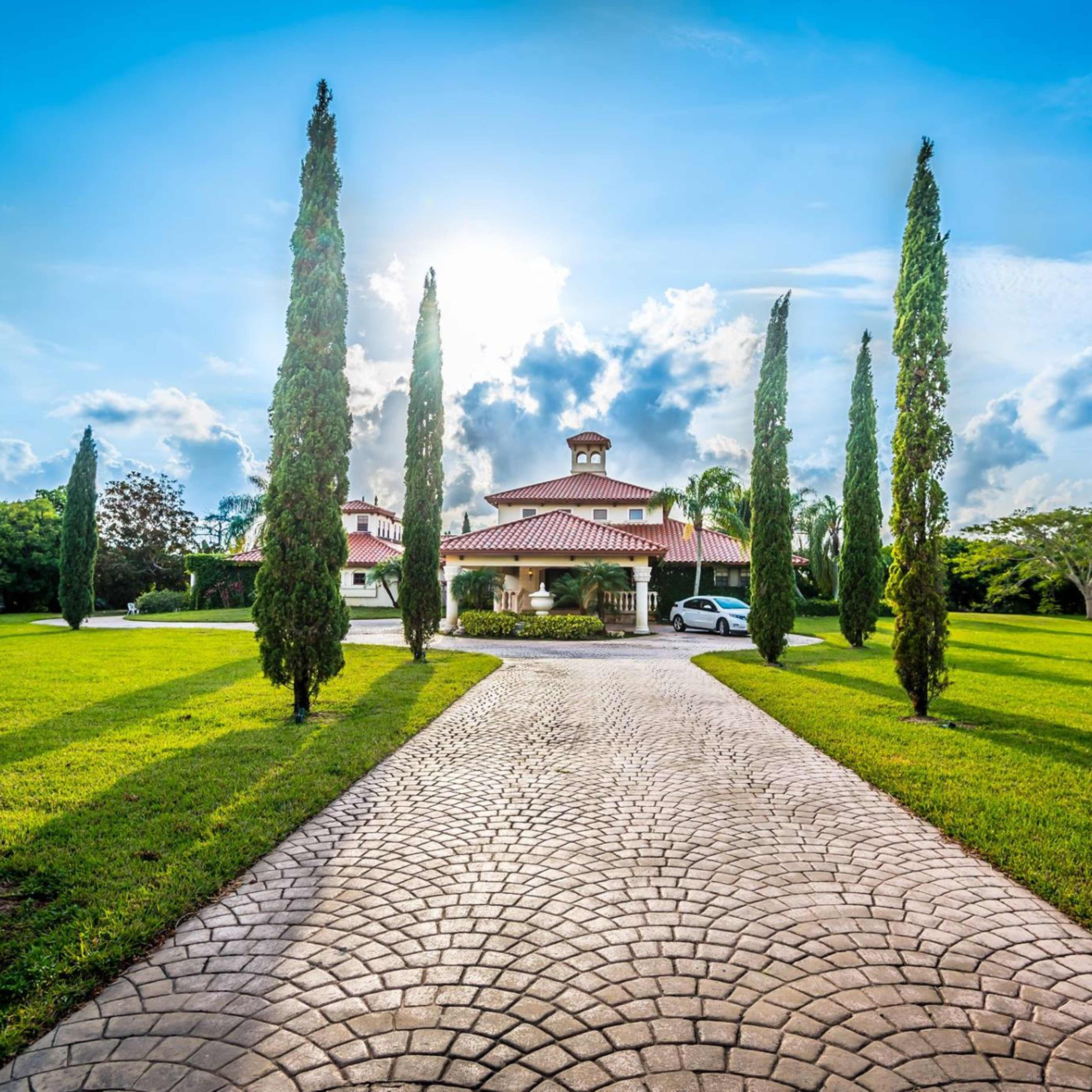 A brick pathway leads to a large house with a red roof, flanked by tall cypress trees and surrounded by a green lawn under a bright sky.