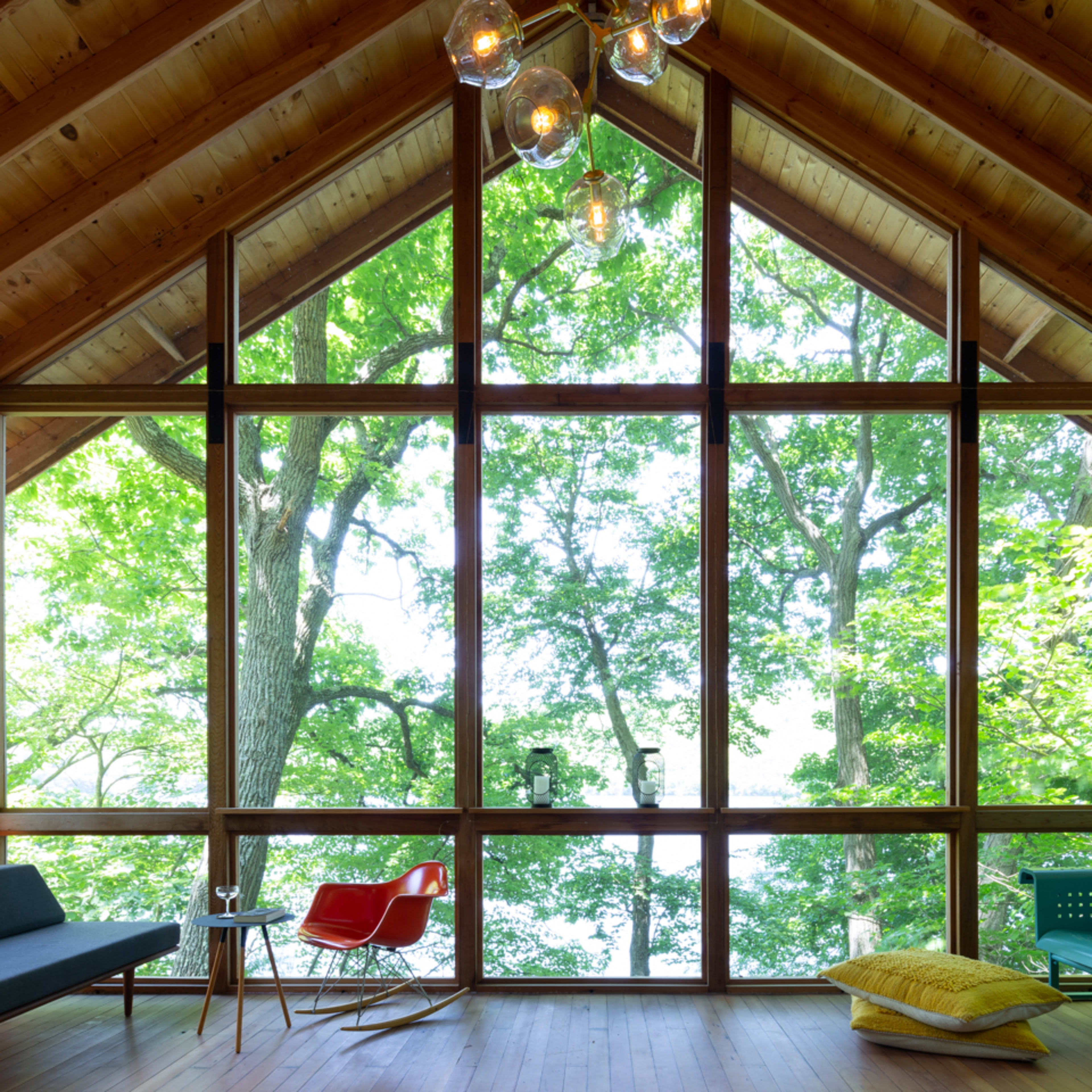 A wooden room with large windows overlooking a lush green landscape, furnished with a black couch, a red chair, and a blue bench.