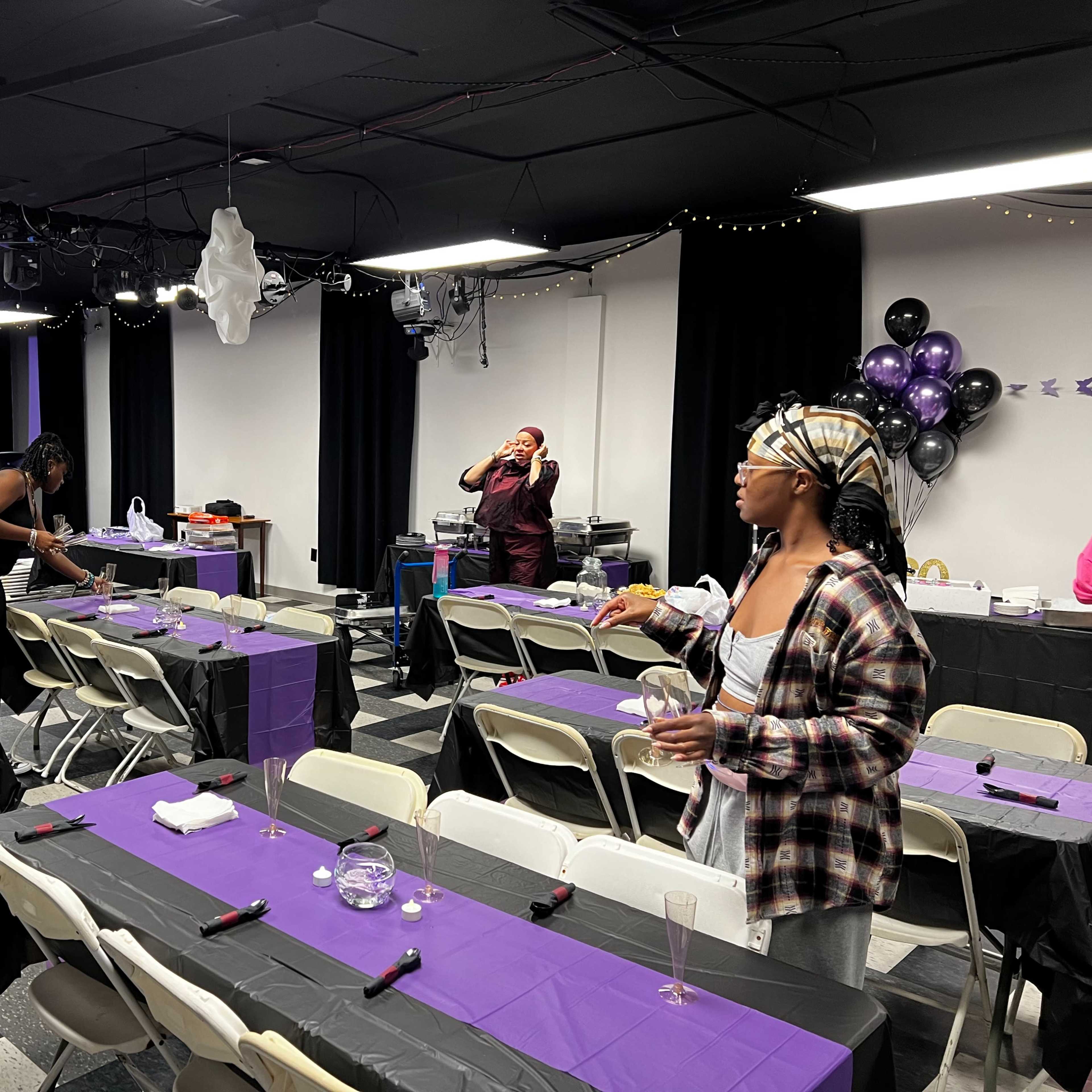 A party setup in a decorated room with purple tables and chairs, a stage area, and balloons, while several people are preparing for the event.