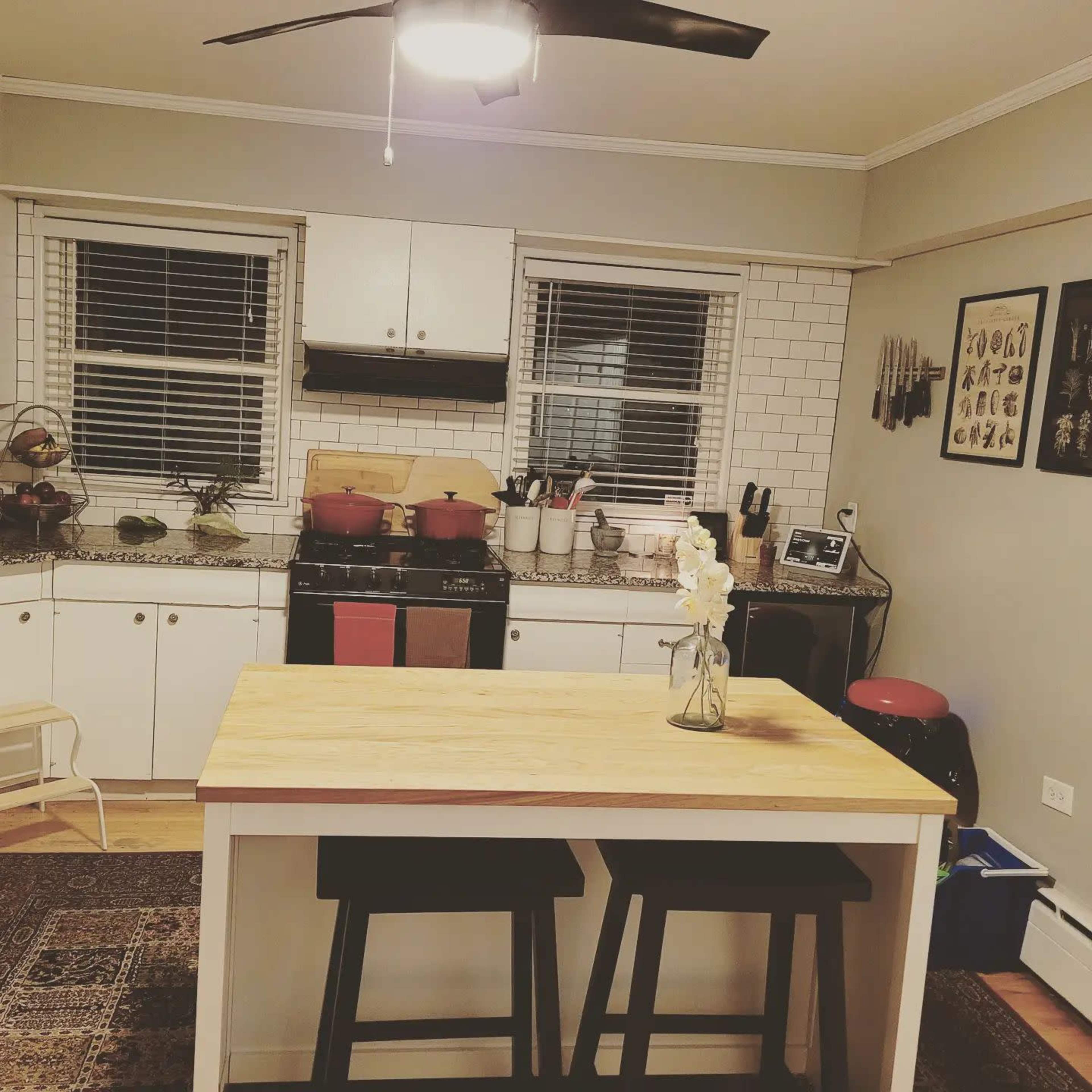 A kitchen with a central wooden table, two black stools, a stove with red pots, and white cabinetry featuring a tiled backsplash.