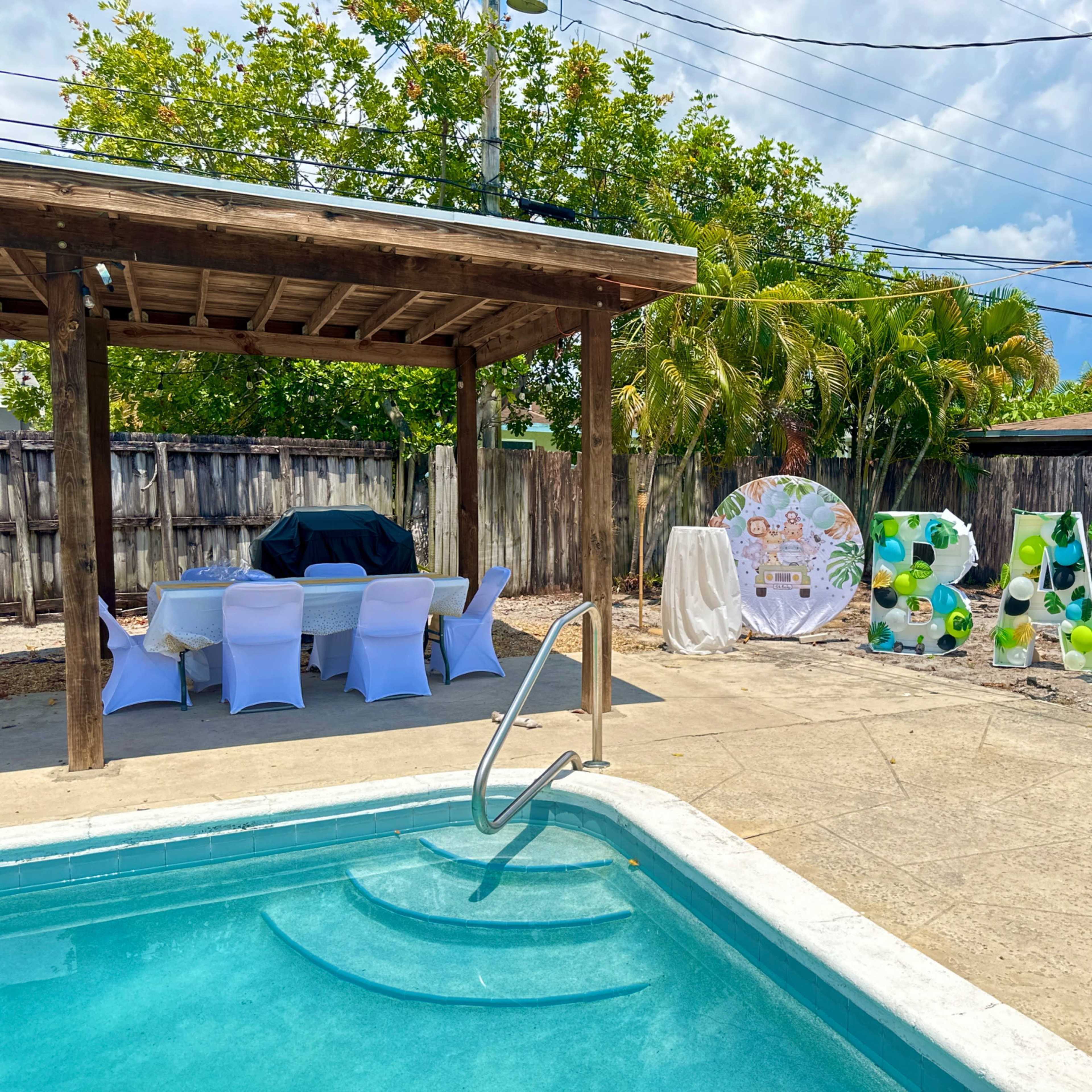 A backyard setting features a swimming pool, a gazebo with tables, and large decorative letters spelling "BABY" surrounded by tropical plants.