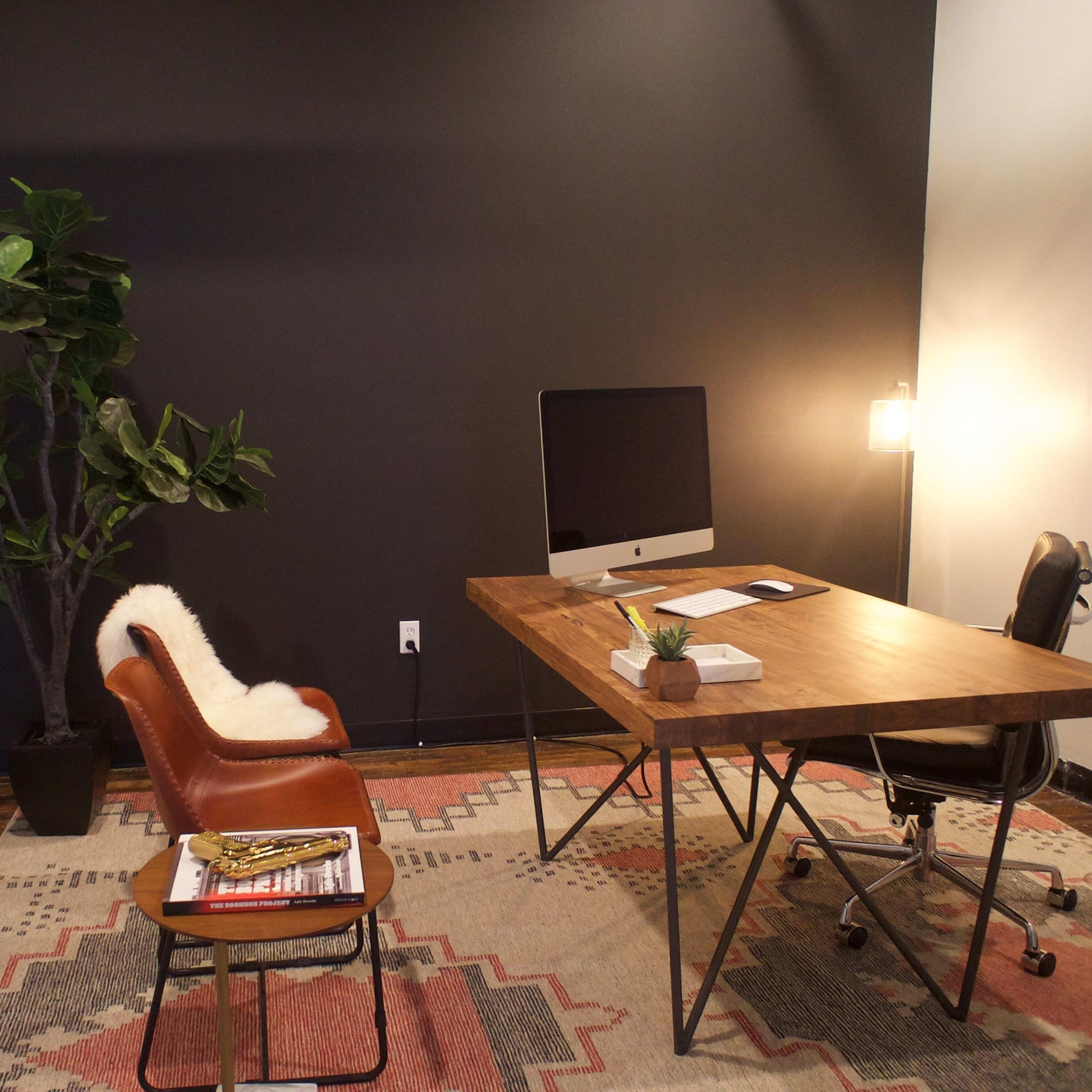 A minimalist office space with a wooden desk, an iMac, a brown leather chair, a potted plant, and a patterned rug on the floor.