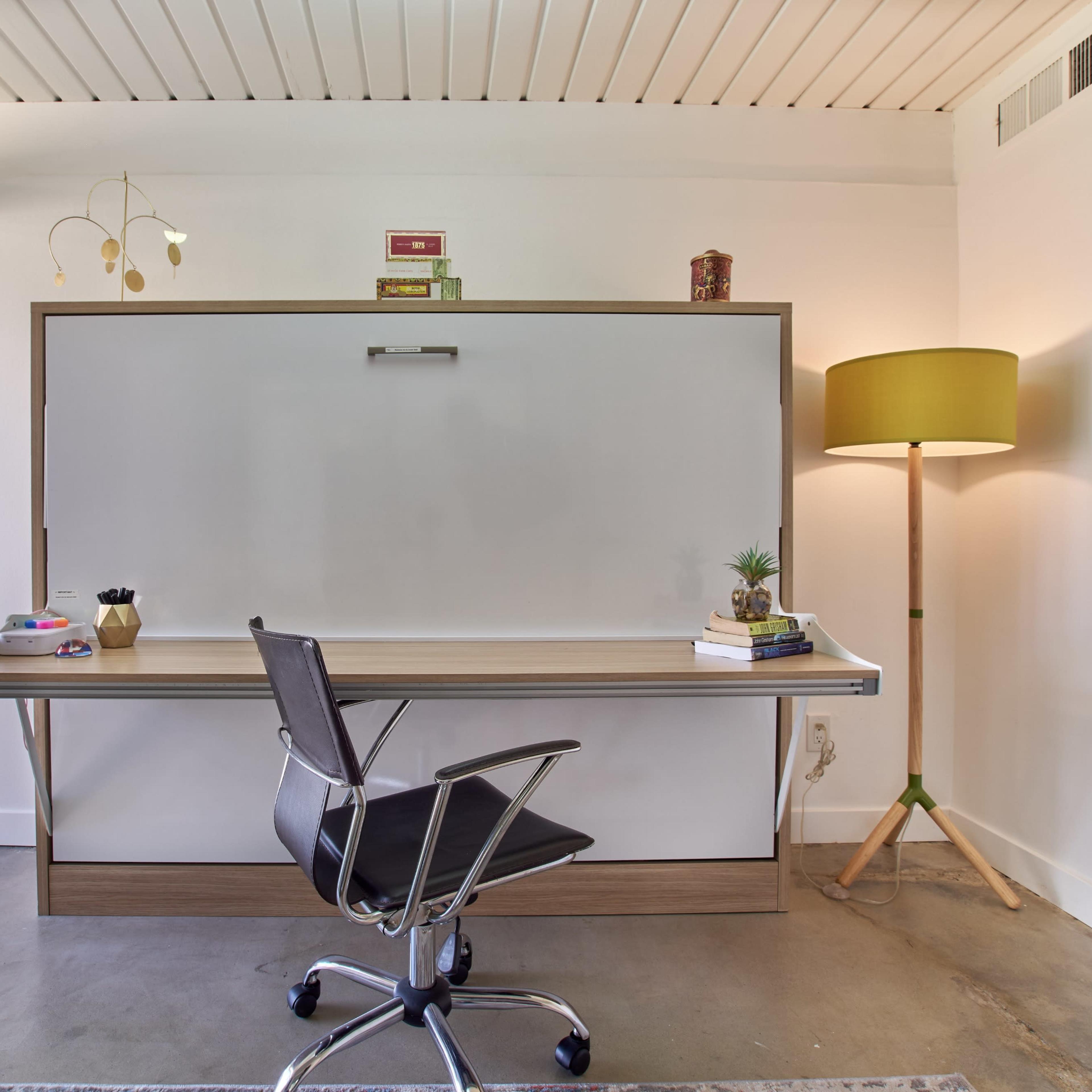 A minimalist home office features a wall-mounted folding desk, a black office chair, and a floor lamp beside a large window.