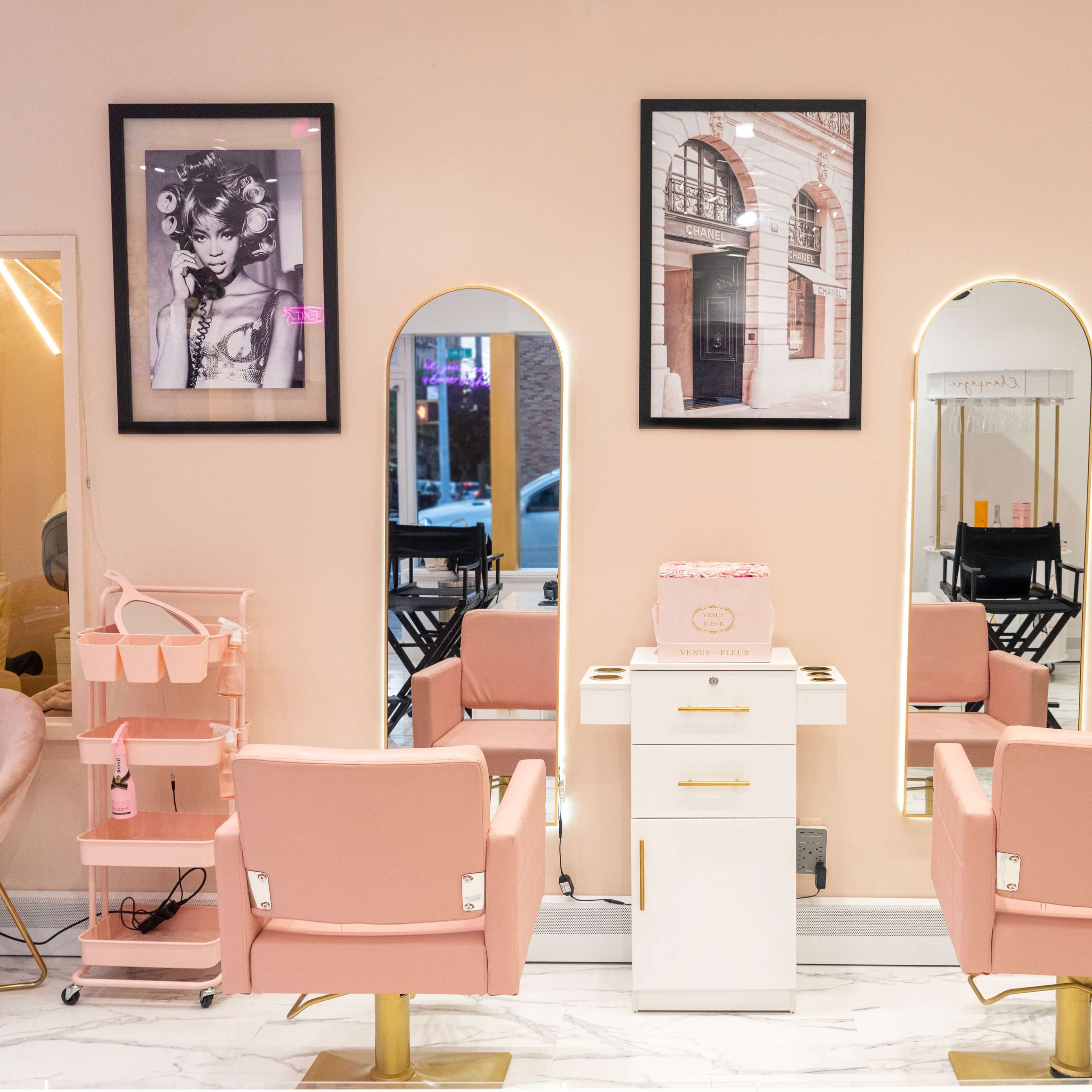 The interior of a hair salon features three mirrors above pink chairs, white styling stations, and framed black-and-white photographs on the walls.