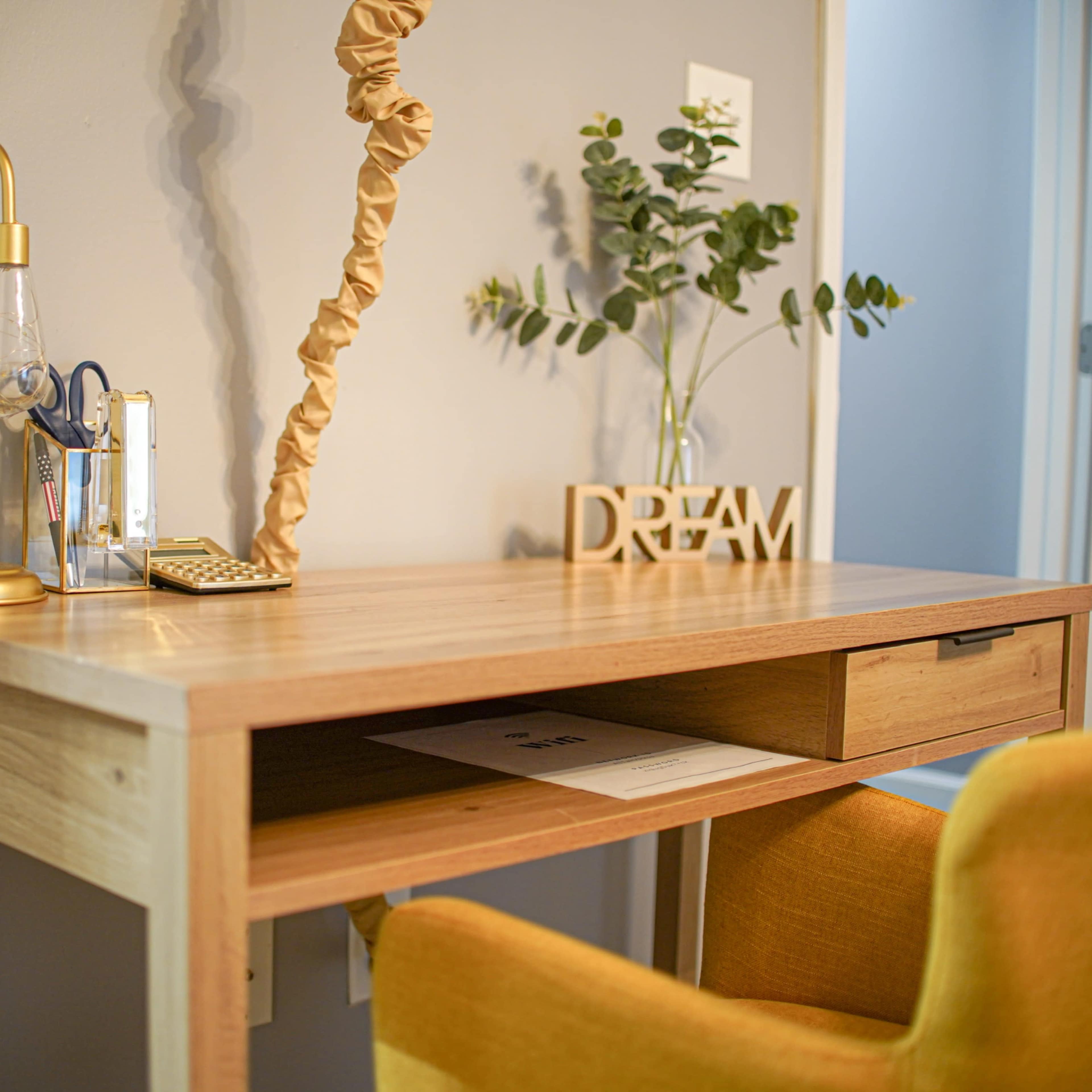 A wooden desk is styled with a lamp, a small plant, and a decorative "DREAM" sign beside a yellow chair.