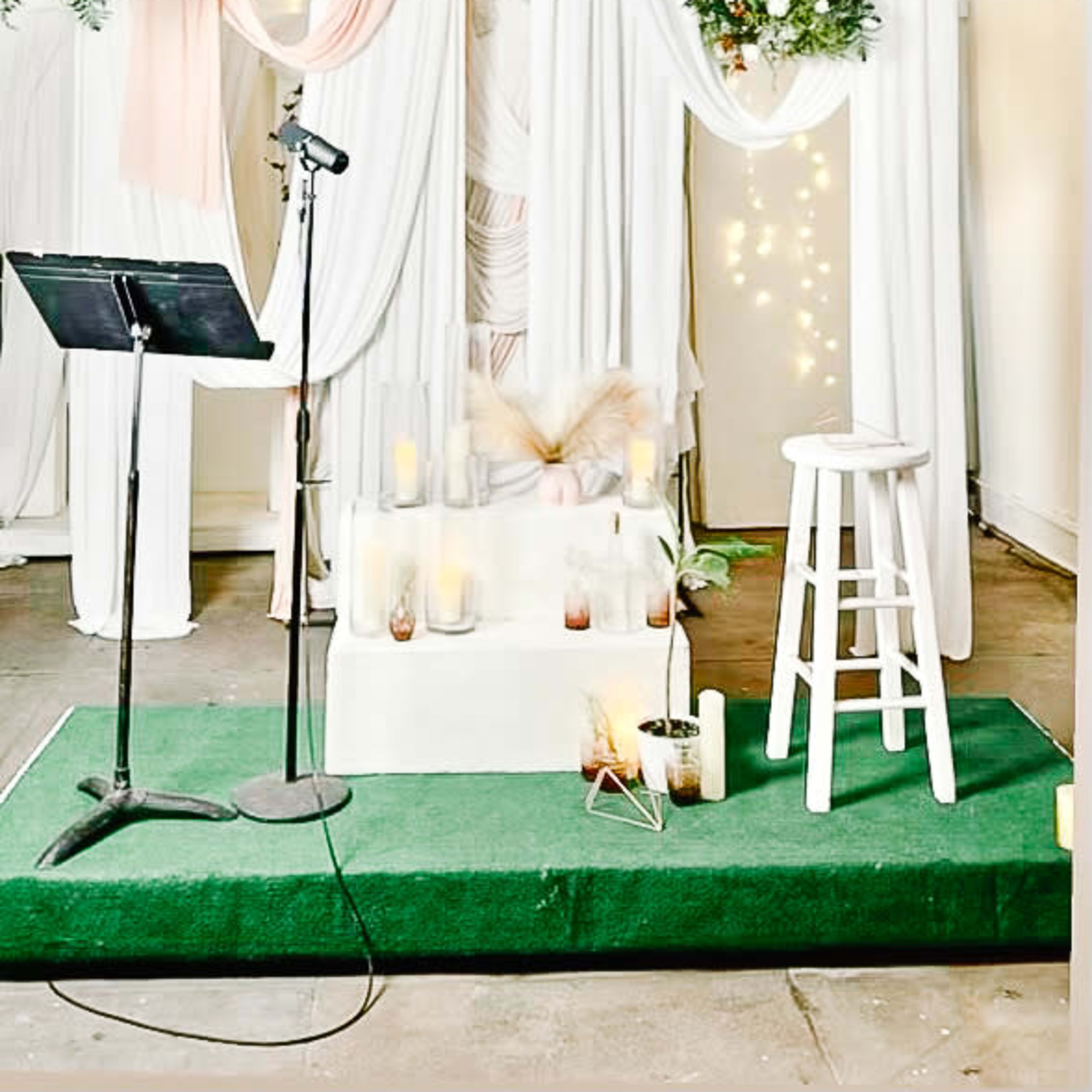 A decorated stage with white drapes, greenery, candles, a microphone stand, and a stool, set up for an event.