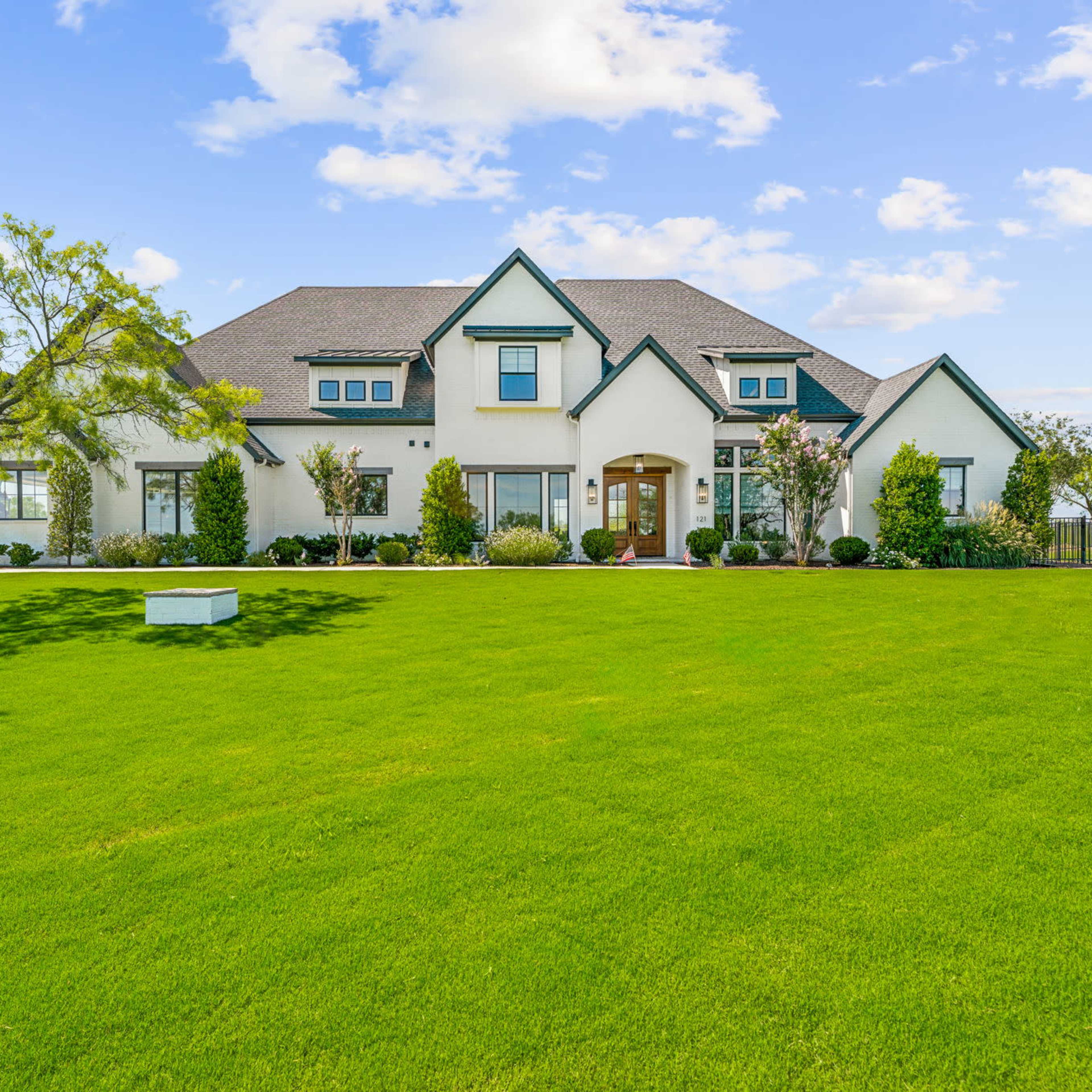 A large two-story house with a landscaped yard and trees is situated on a green lawn under a blue sky.