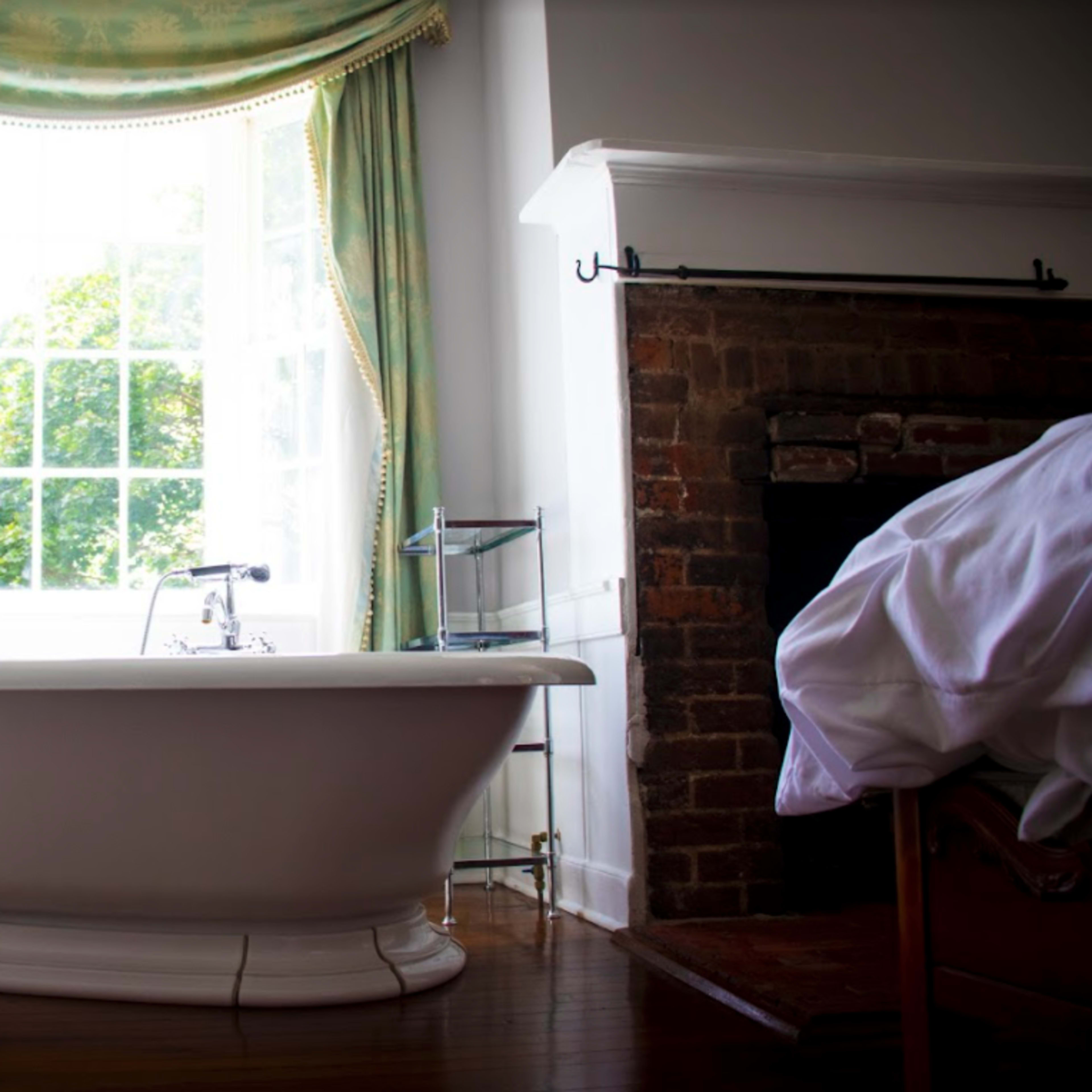 A freestanding bathtub is positioned near a window with green curtains, next to a brick fireplace in a well-lit room.