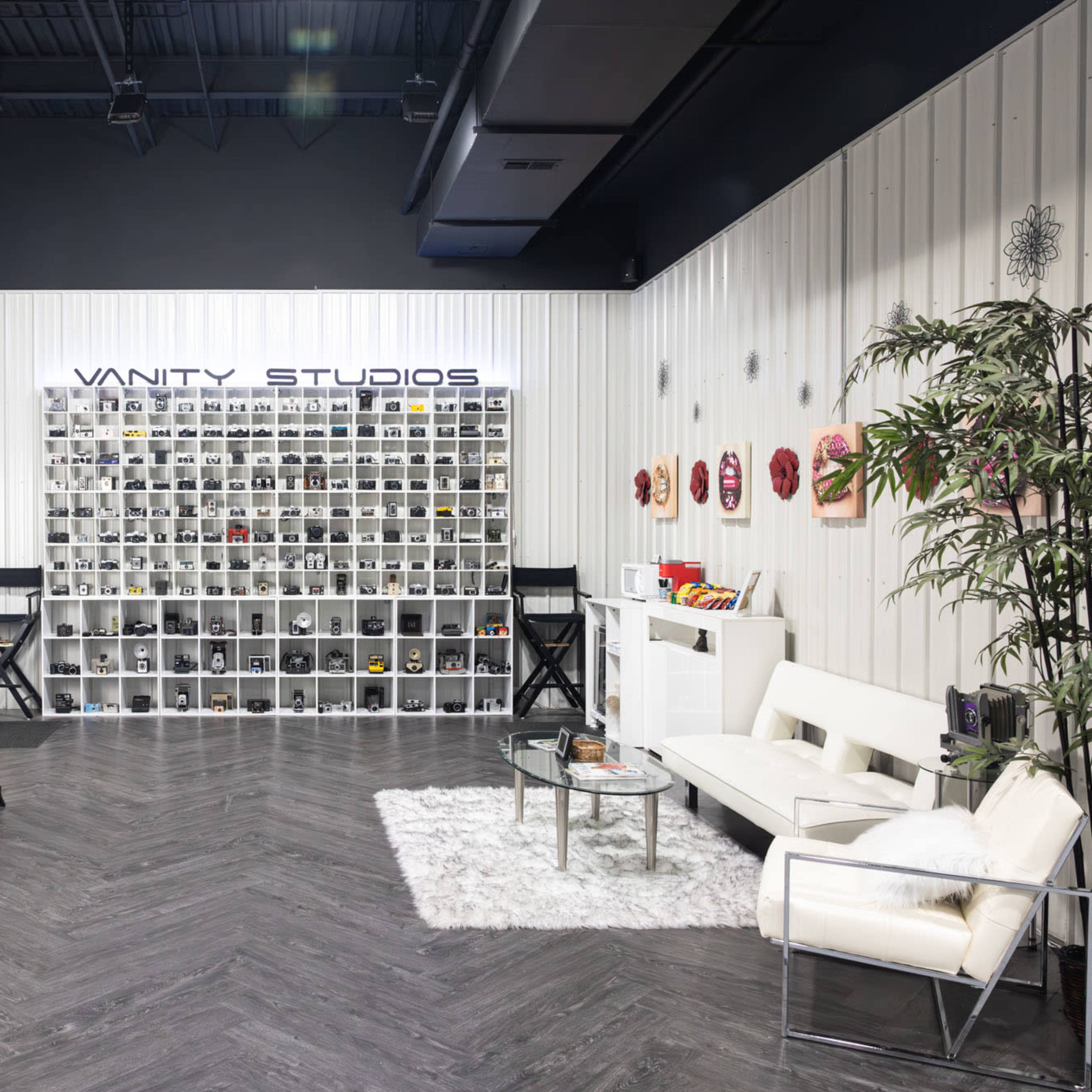 A spacious studio interior with a makeup vanity area, shelves filled with various products, and a seating area featuring a white sofa and a glass coffee table.