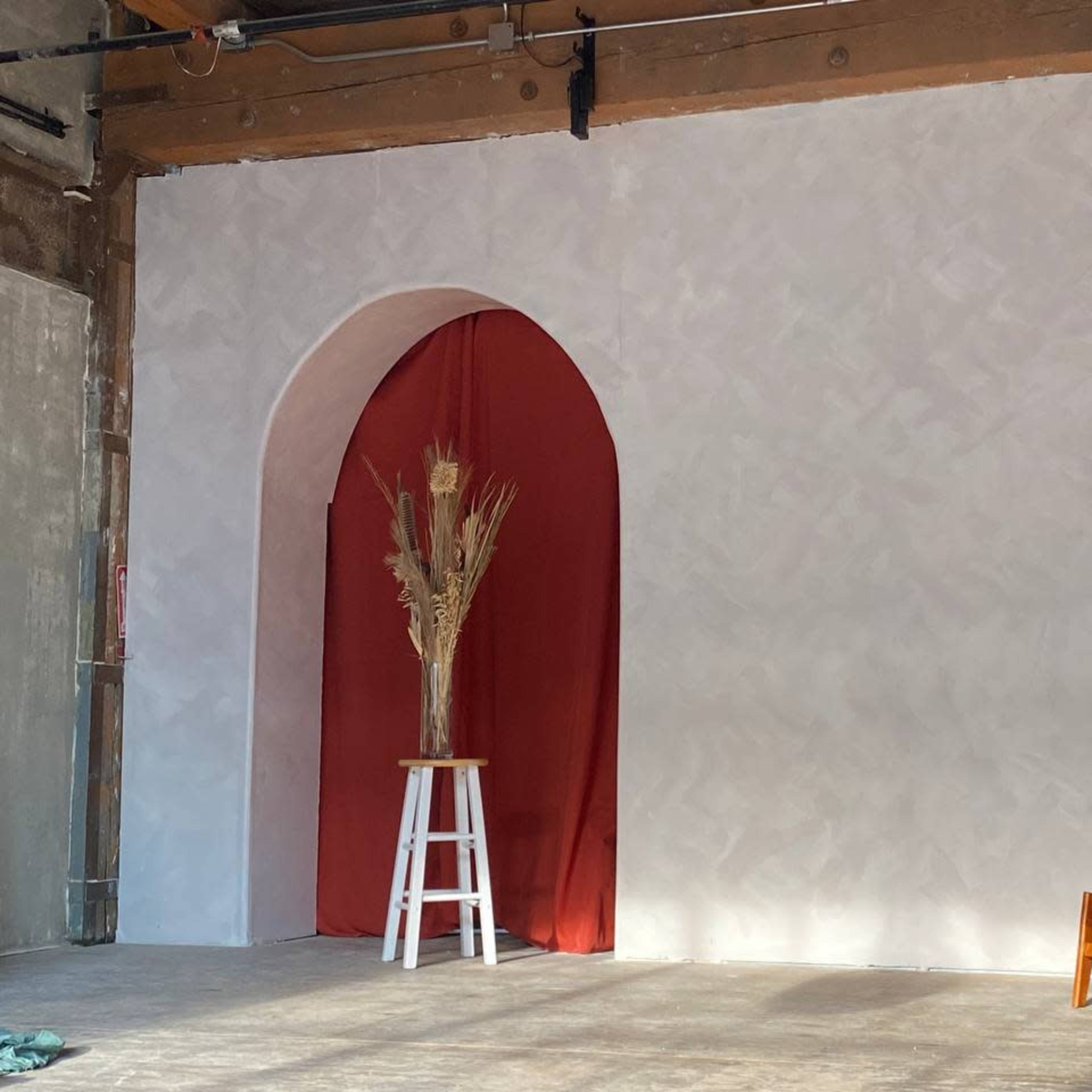 A minimalist interior space featuring a white wall with an archway covered in red fabric, a tall vase with dried plants, and a wooden chair beside a white stool.