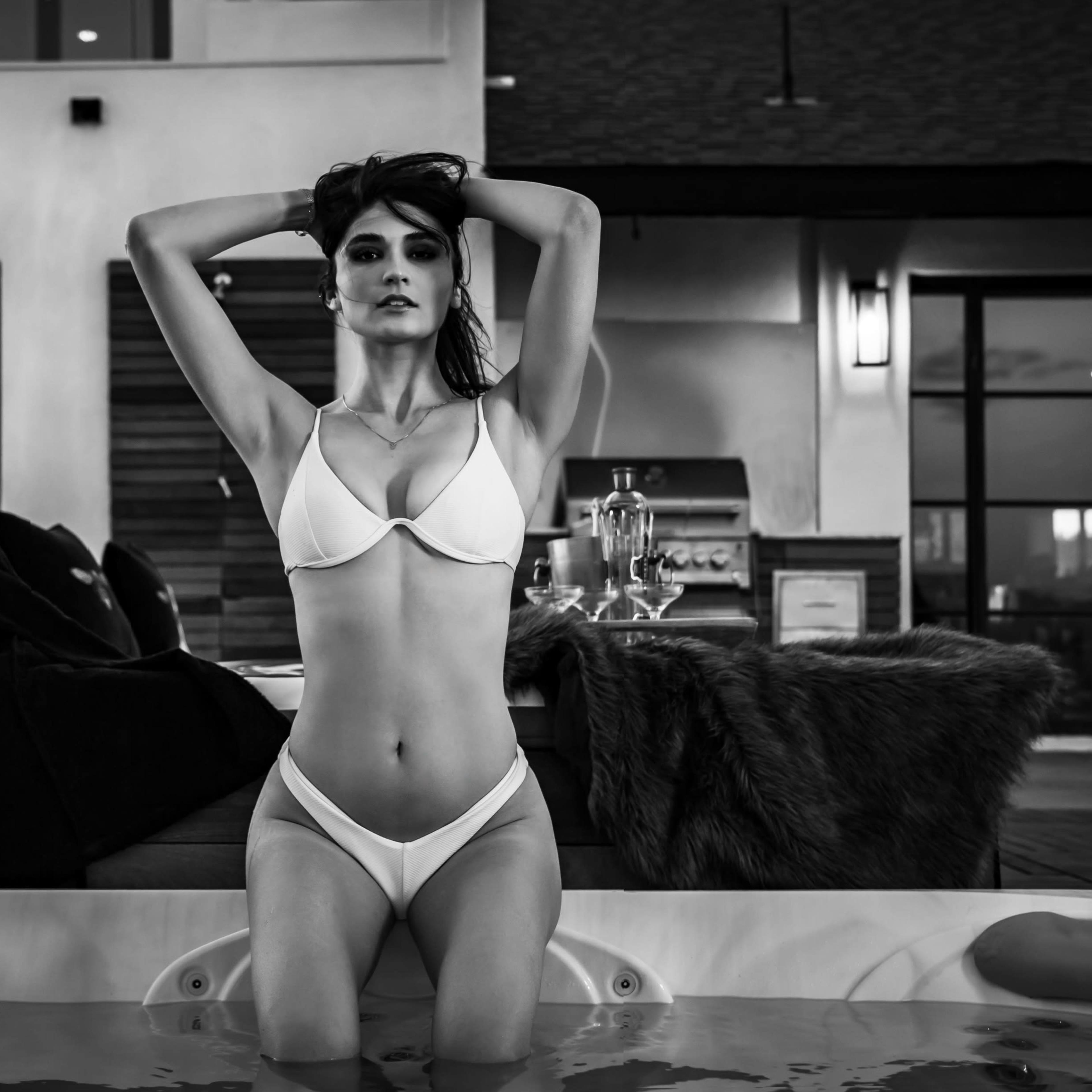 A woman in a white bikini poses in a hot tub with her hands behind her head, seated next to a fur throw and a small table with a drink.