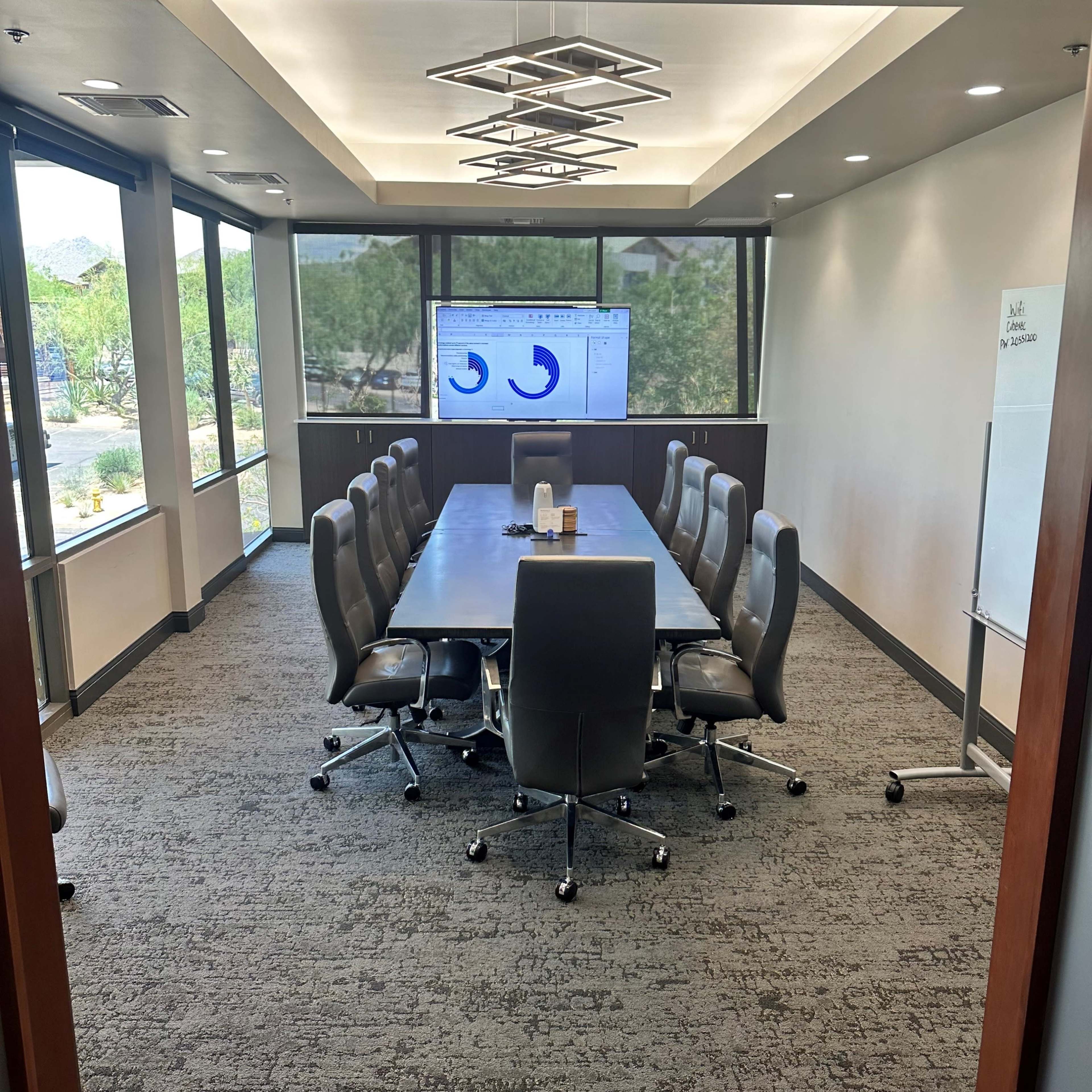 A modern conference room features a long table surrounded by ergonomic chairs, with a large screen displaying a graph and windows that provide natural light.